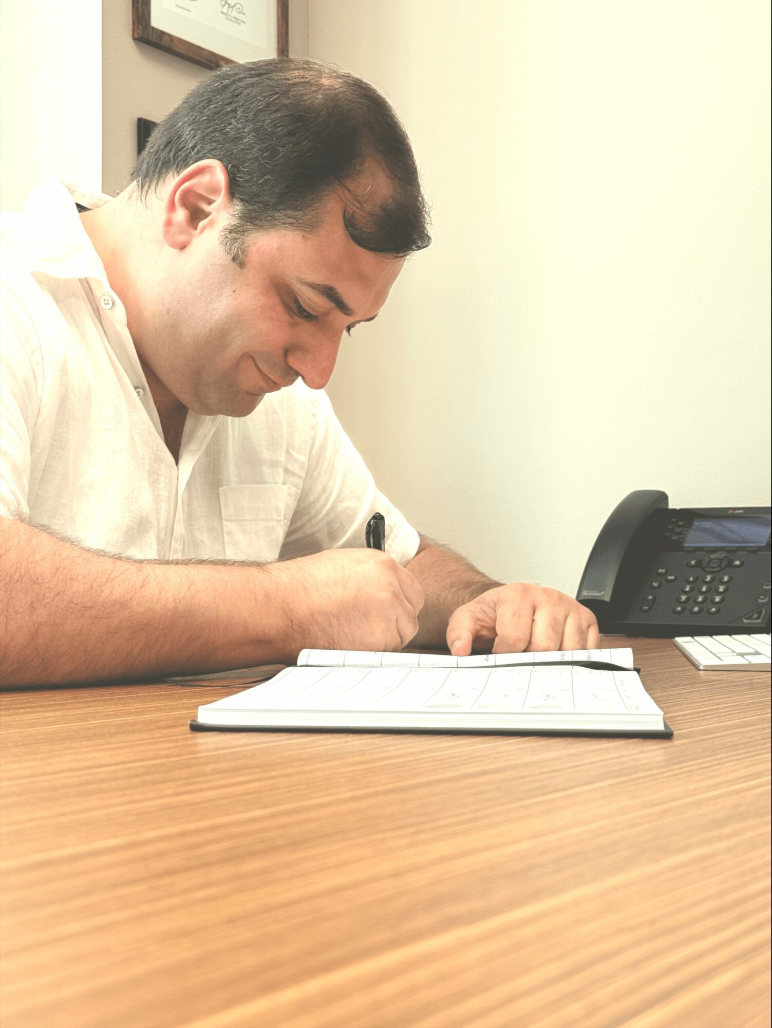 Person writing in a notebook at a desk with a phone nearby, smiling and focused.