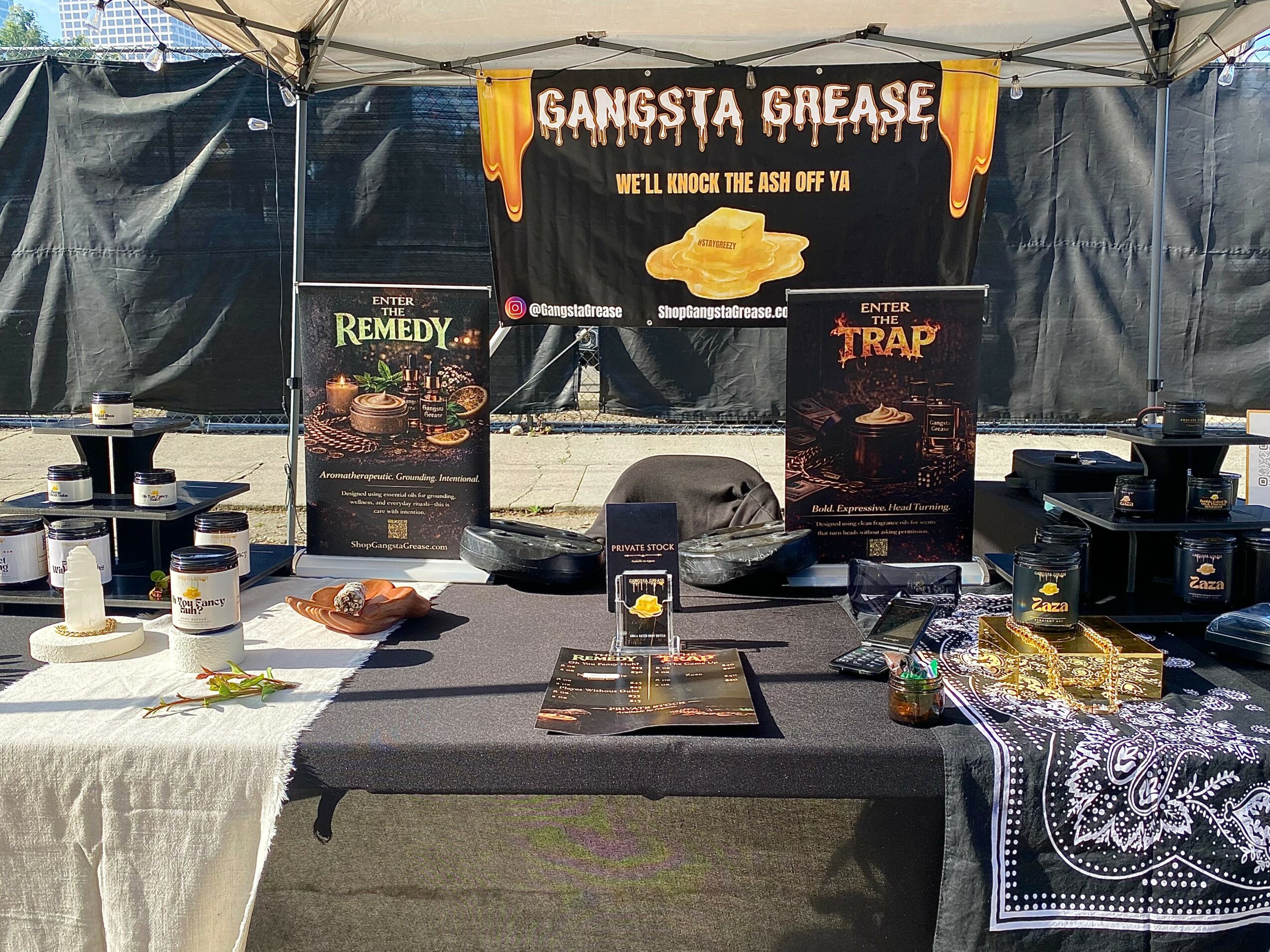 Booth with banners reading 'Gangsta Grease,' 'Remedy,' and 'Trap,' displaying food and promotional materials, under a canopy.