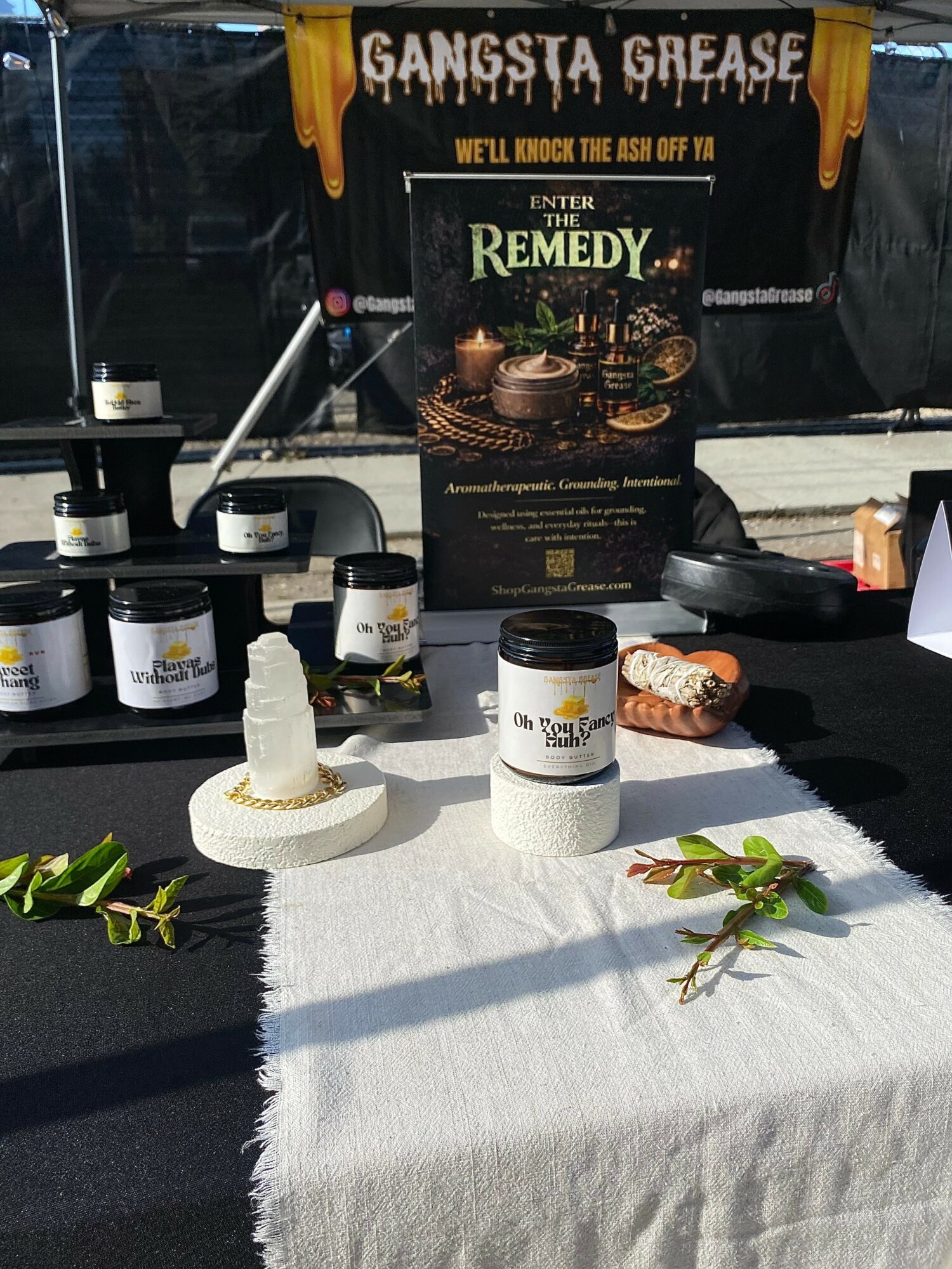 Display table with jars, a sign, and decorative plants outdoors, sunlight casting shadows, with a black banner in the background.