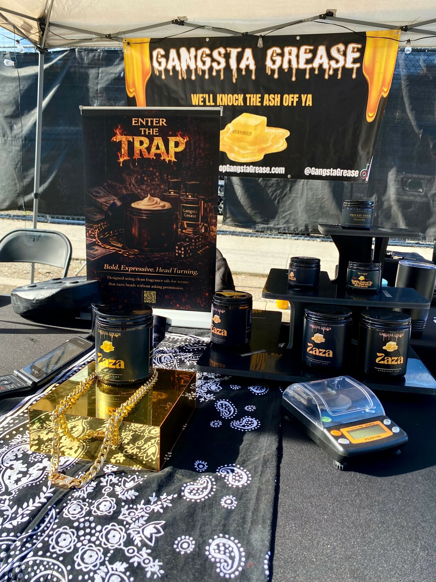Table with black jars, a gold chain, and a promotional sign for Gangsta Grease and Enter the Trap at an outdoor event.