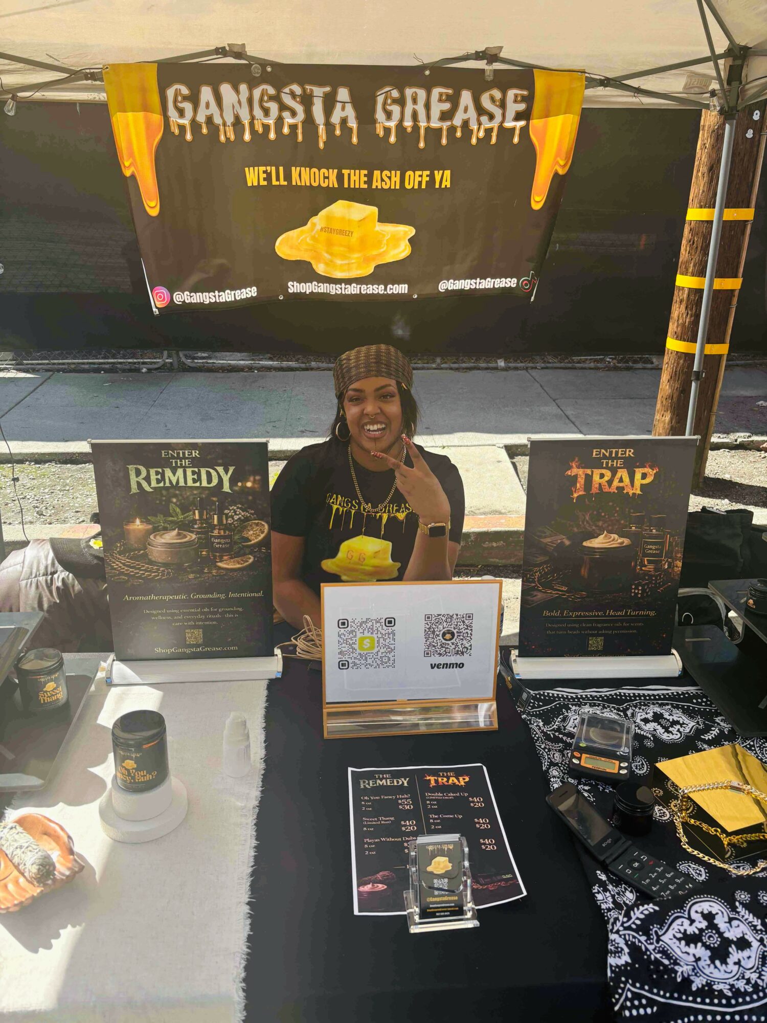 Smiling woman standing behind a table with promotional posters and items, under a tent with a 'Gangsta Grease' banner.