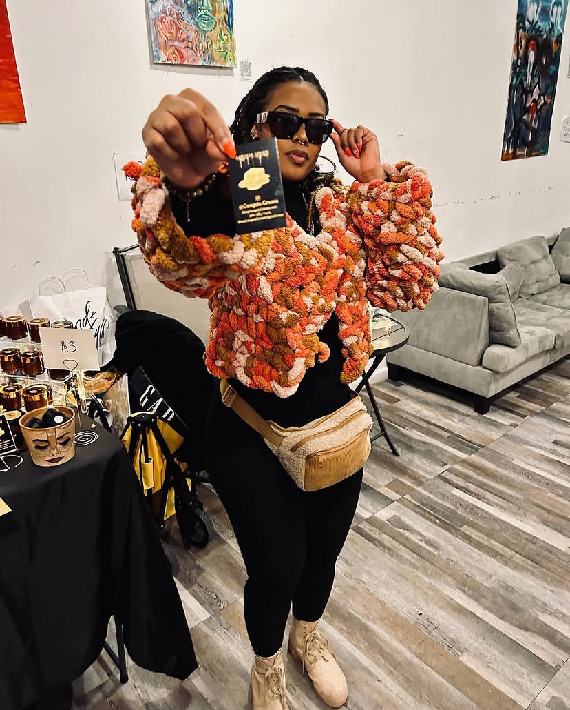 Person taking a selfie in a room with artwork, wearing sunglasses, a colorful textured jacket, black pants, and beige shoes.