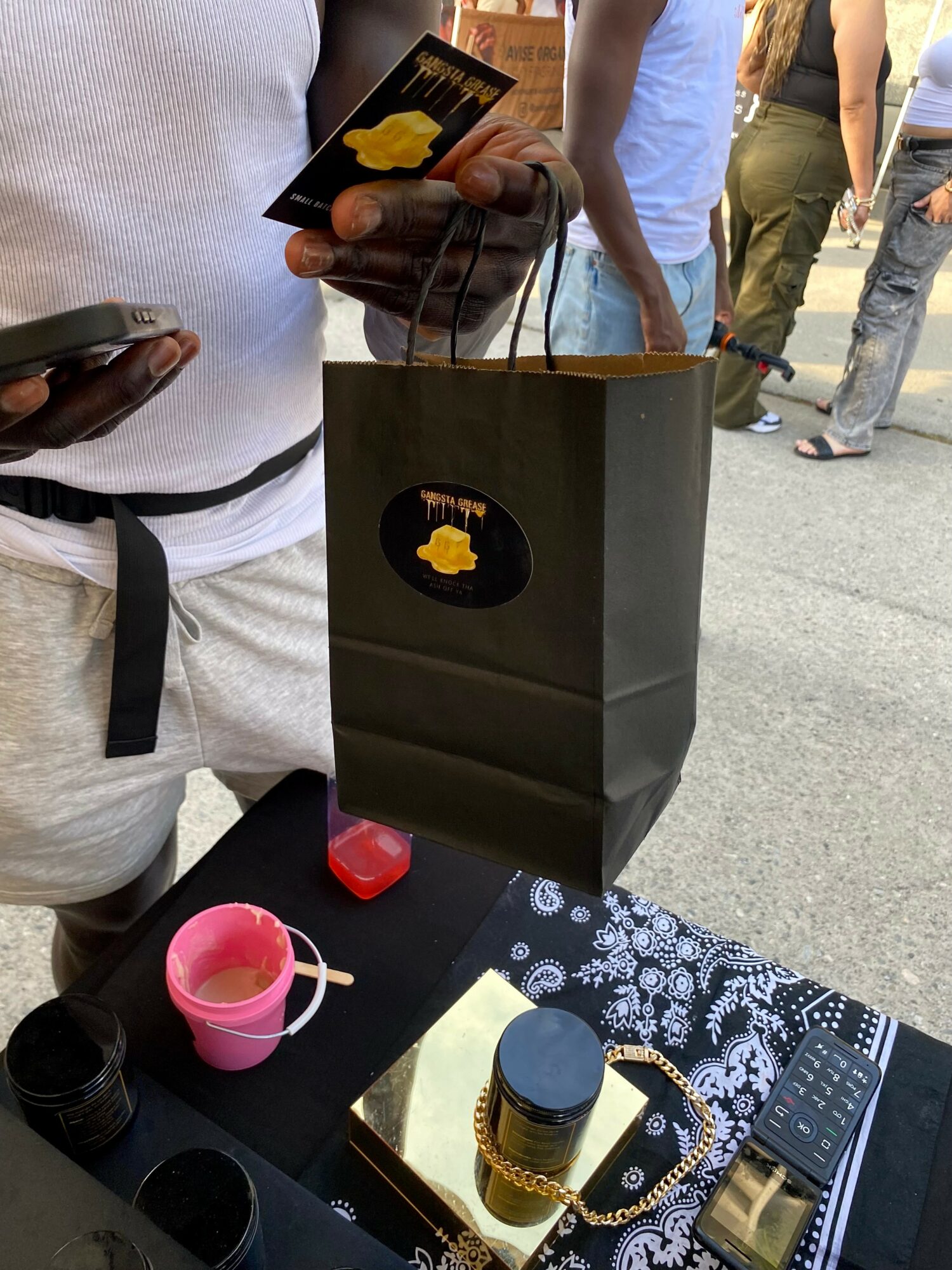 Person holding a black shopping bag with a yellow logo, standing outdoors with people in the background.