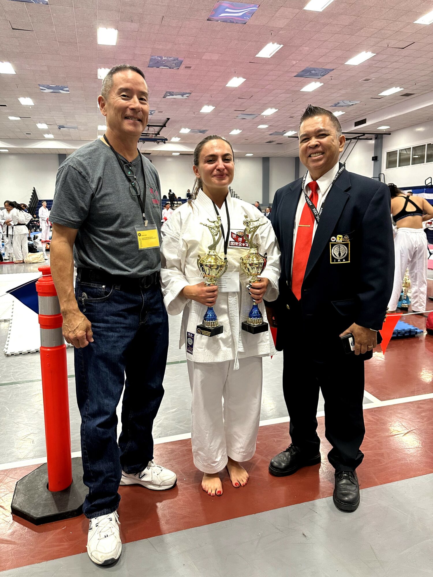 Three people standing indoors, one in a martial arts uniform holding trophies, two men in casual and formal attire, smiling.