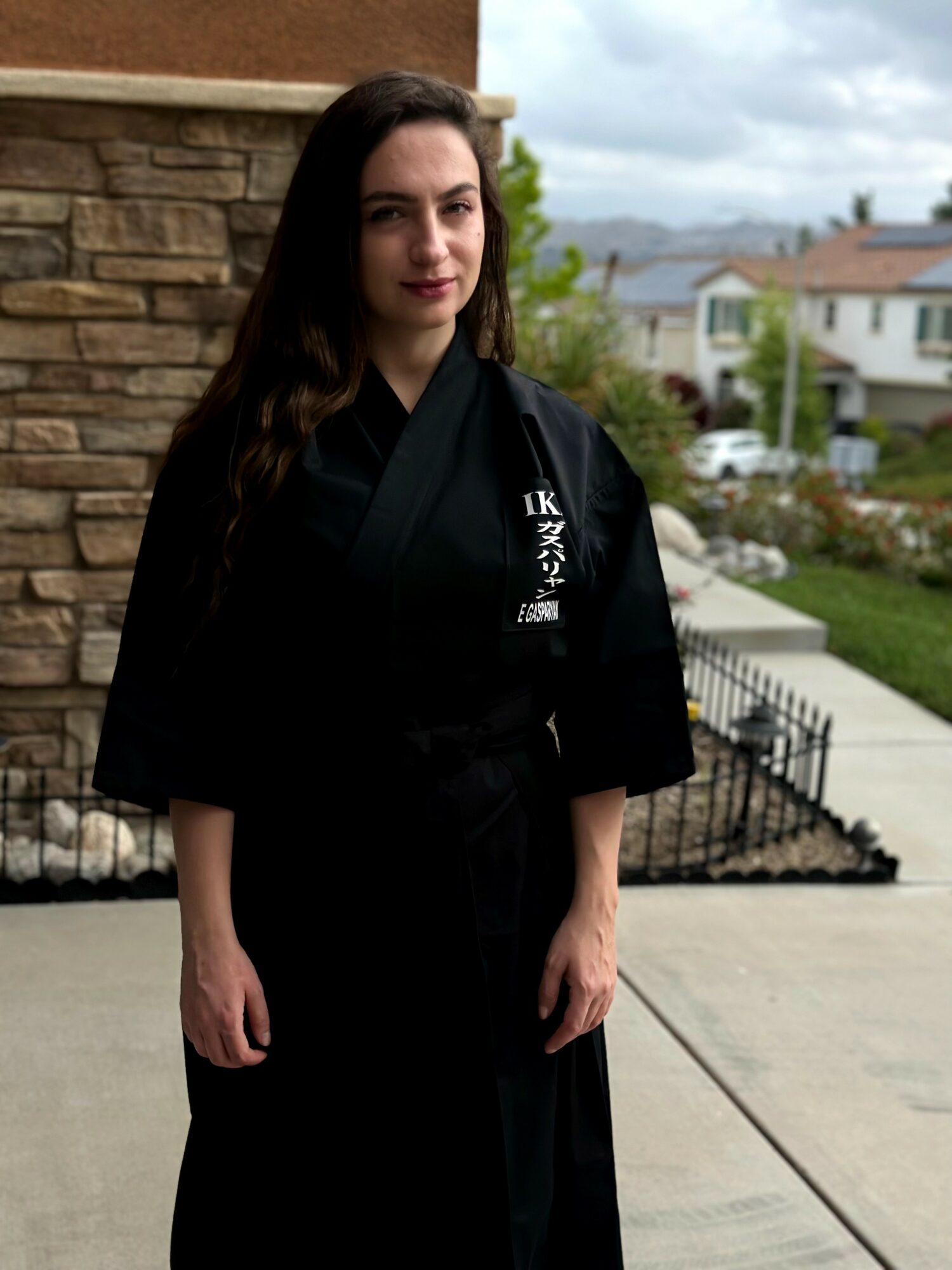 Young woman in a black martial arts uniform standing outdoors near a stone wall, with houses and trees in the background.