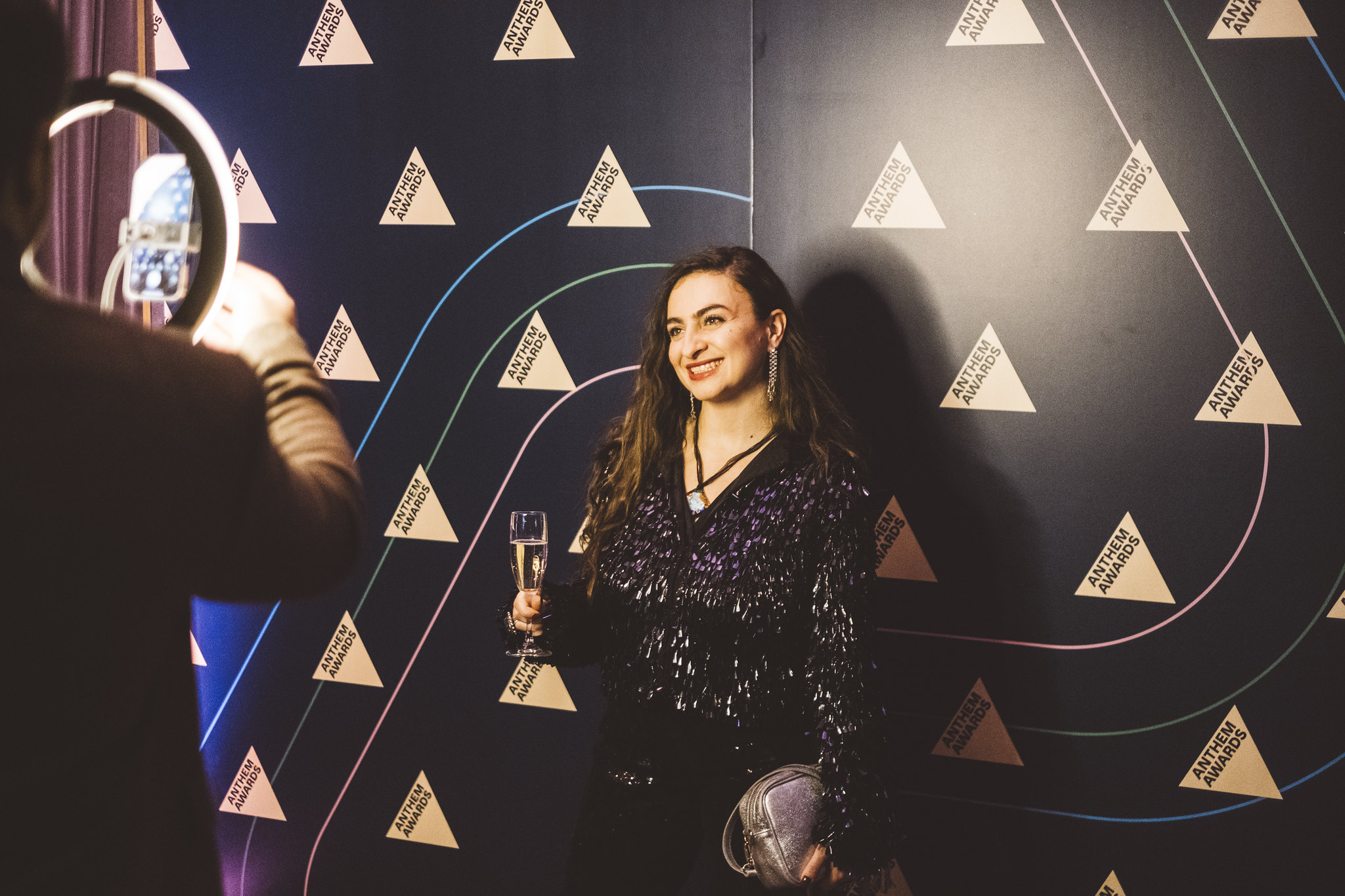 Woman holding a glass, smiling, in front of a dark patterned background with geometric shapes.
