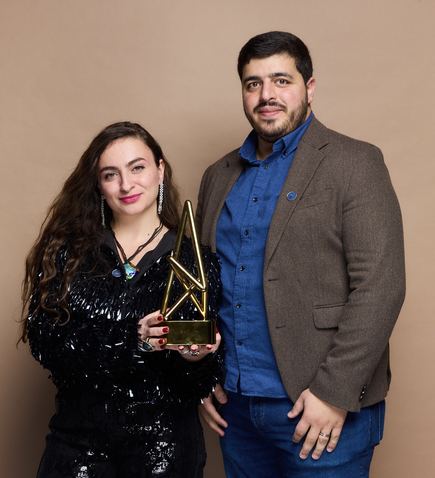 A woman and a man stand together holding a geometric trophy, both smiling, against a plain beige background.