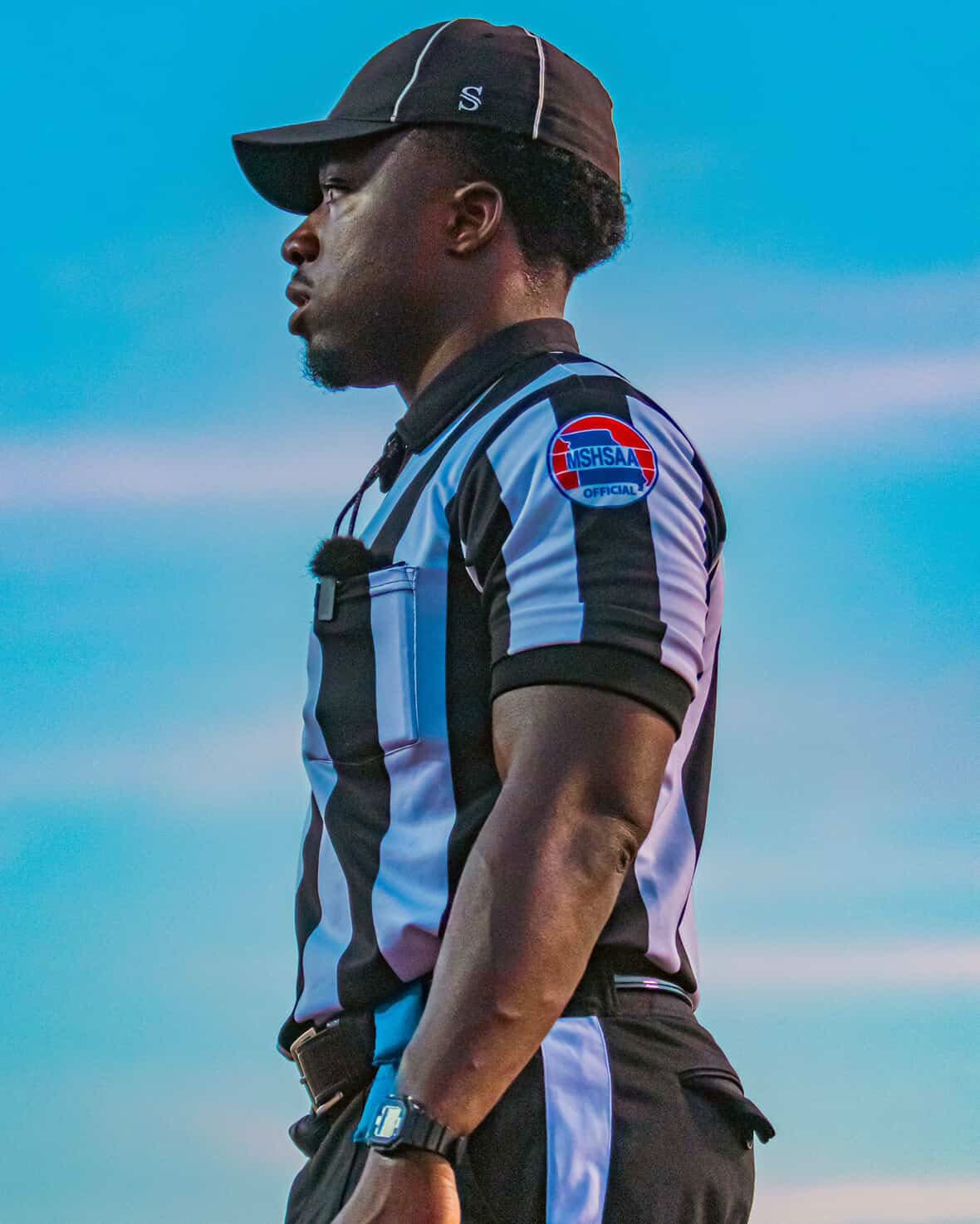 Side view of a referee in a black cap and striped shirt against a blue sky.