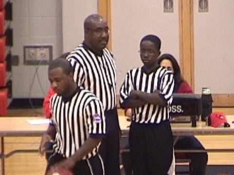 Three referees in black-and-white striped shirts standing in a room with a woman and a desk in the background.
