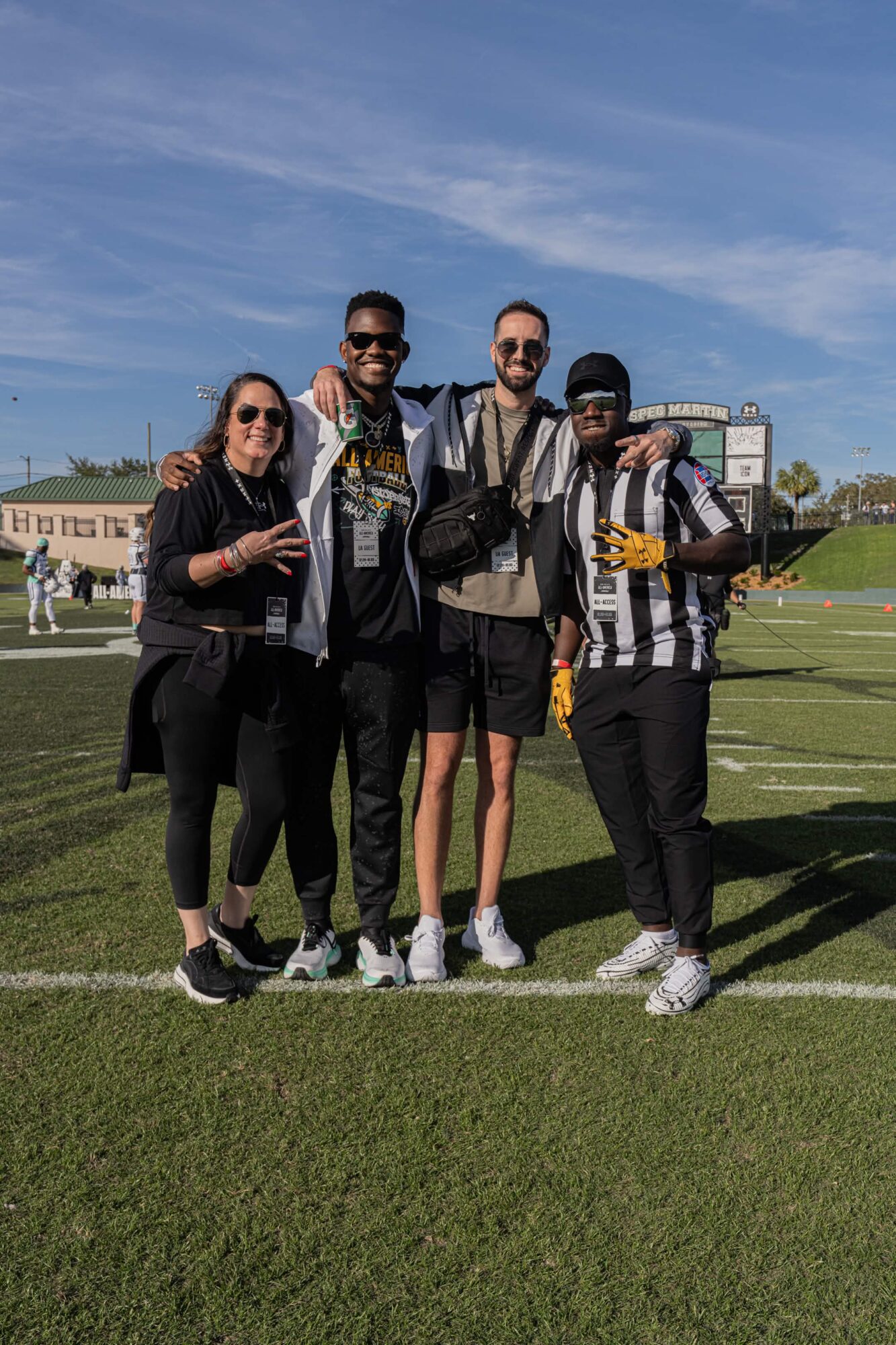 Four people standing on a football field, smiling, with a blue sky and stadium in background.
