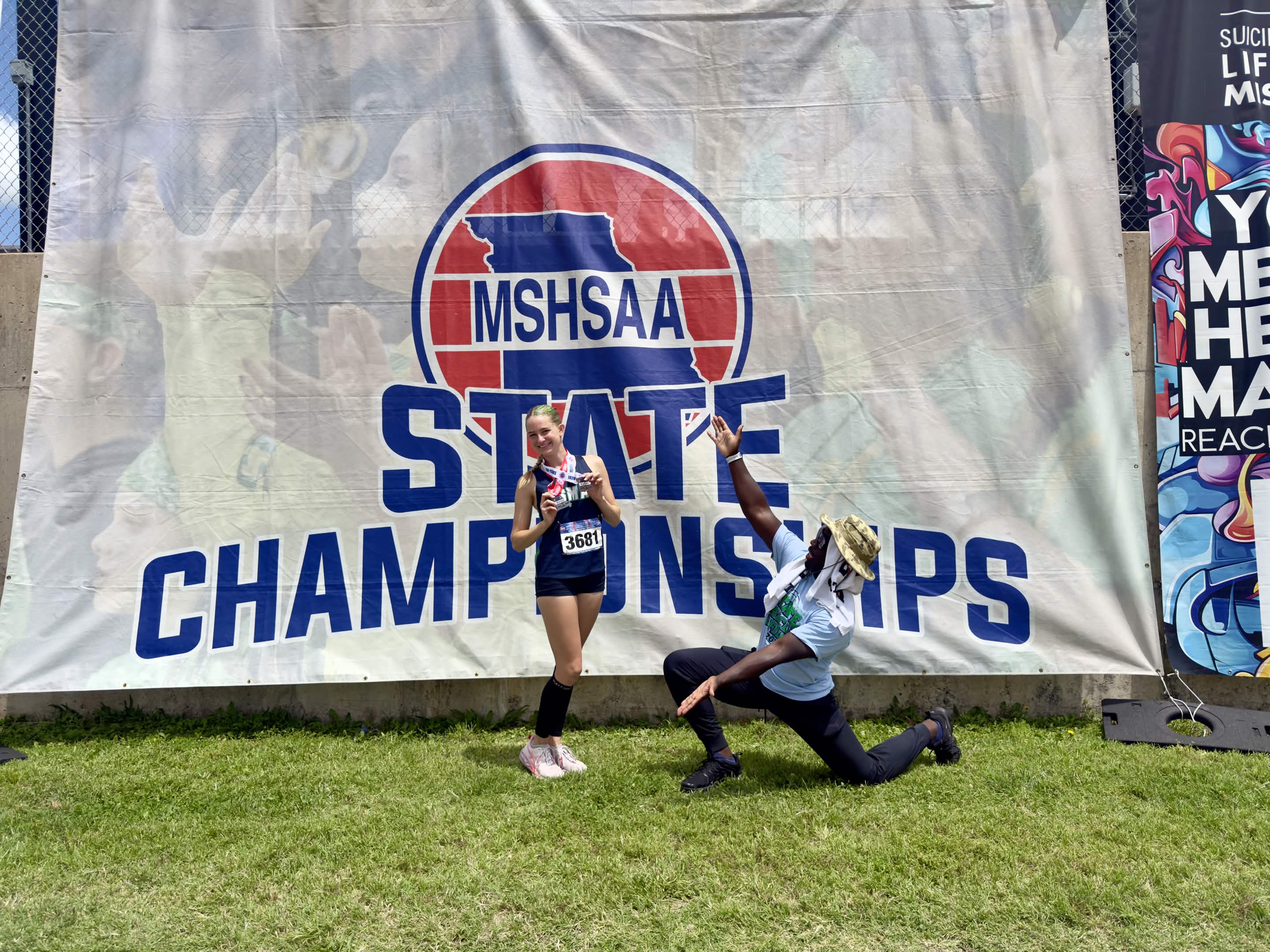 Two people in front of a large banner for MSHSAA State Championships, one kneeling and one standing, on grass.