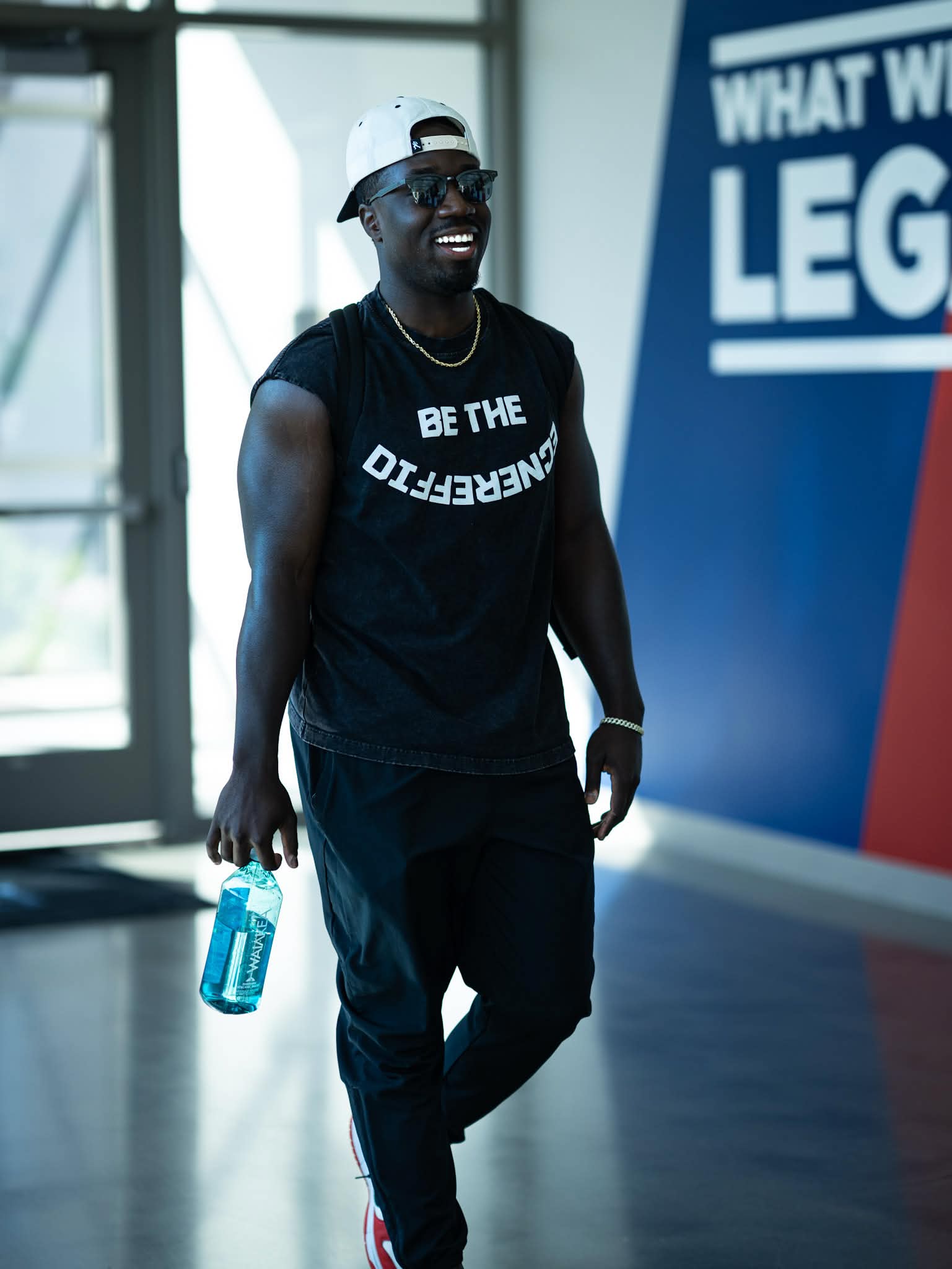 Smiling man wearing sunglasses, a cap, and a sleeveless shirt holding a water bottle indoors.