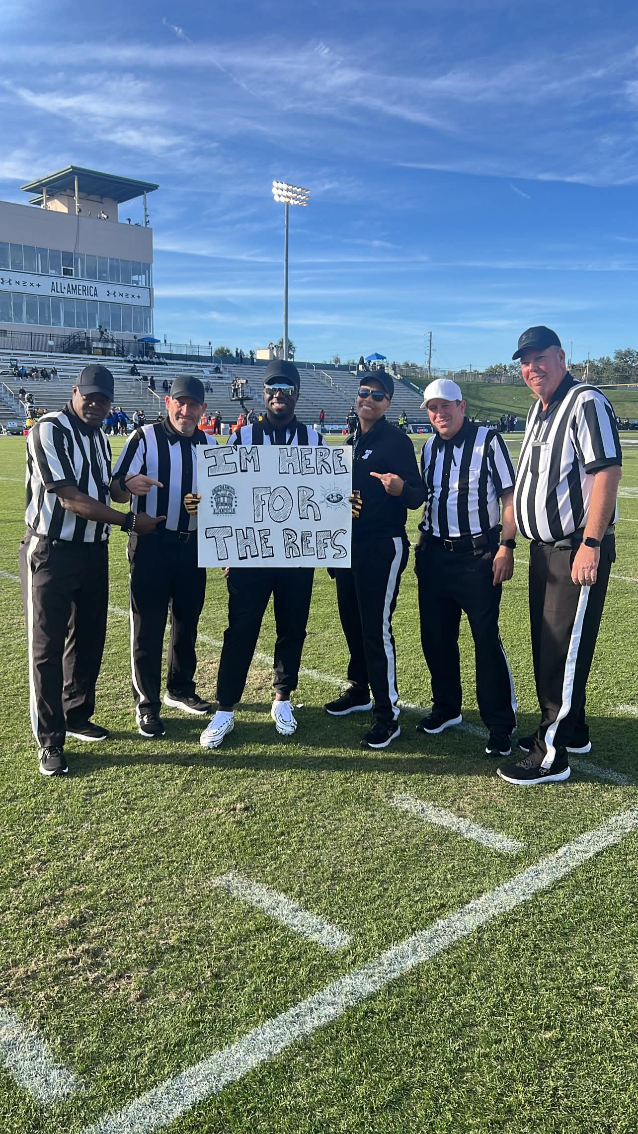 Group of six referees holding a sign on a football field with a stadium and blue sky in background.