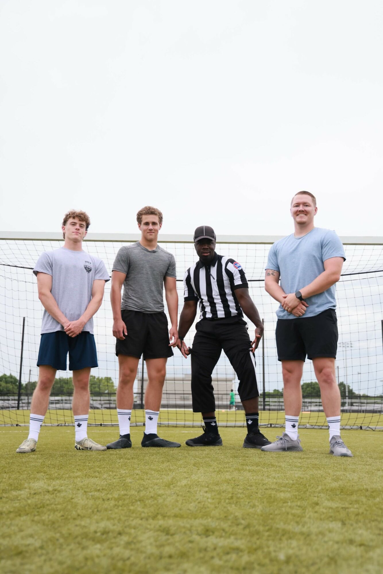 Four people standing on a sports field, with a fence and sky in the background, smiling and posing for the photo.