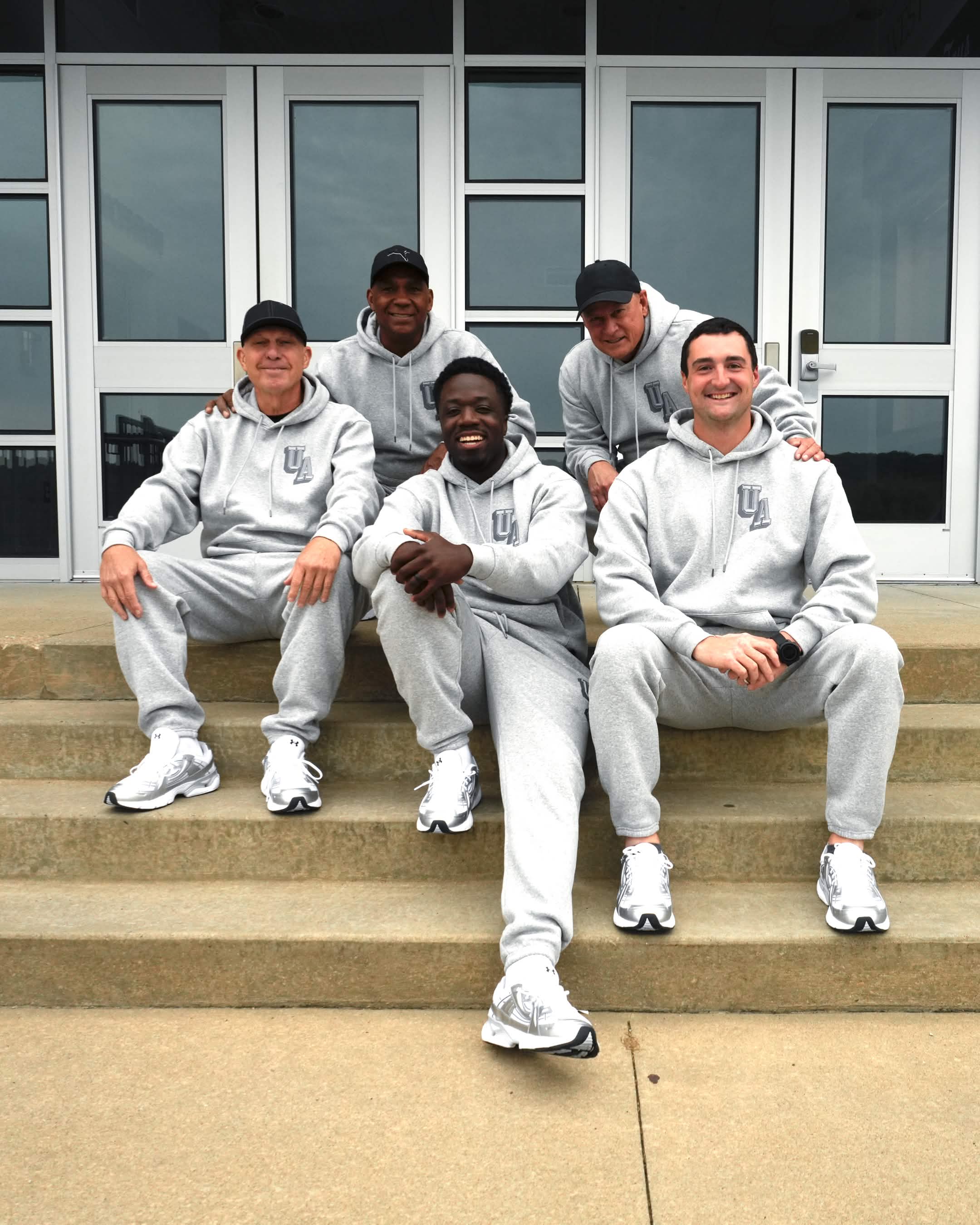 Five people sitting on steps outside a building, wearing matching light gray outfits and caps, smiling at the camera.
