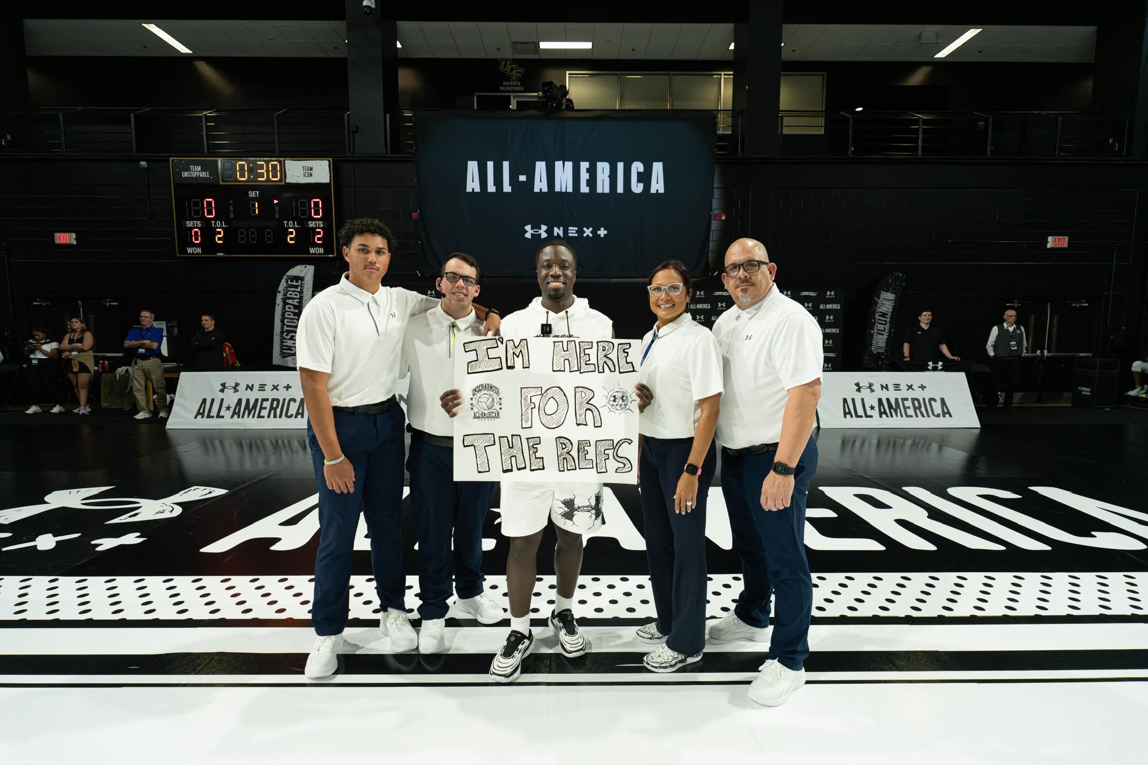Group of five people standing on a basketball court holding a sign that says 'SM HERE FOR THE BEARS' with a large screen behind them displaying 'ALL-AMERICA'.