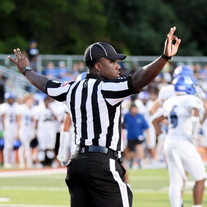 Referee signaling a call during a football game with players and spectators in the background.