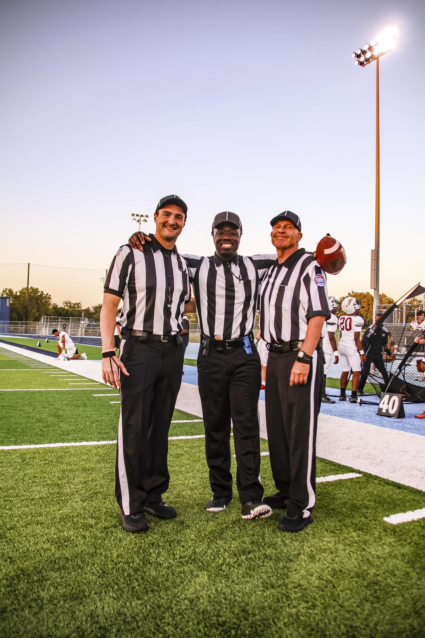 Three referees in black and white striped shirts standing on a football field, smiling, with a stadium and players in the background.