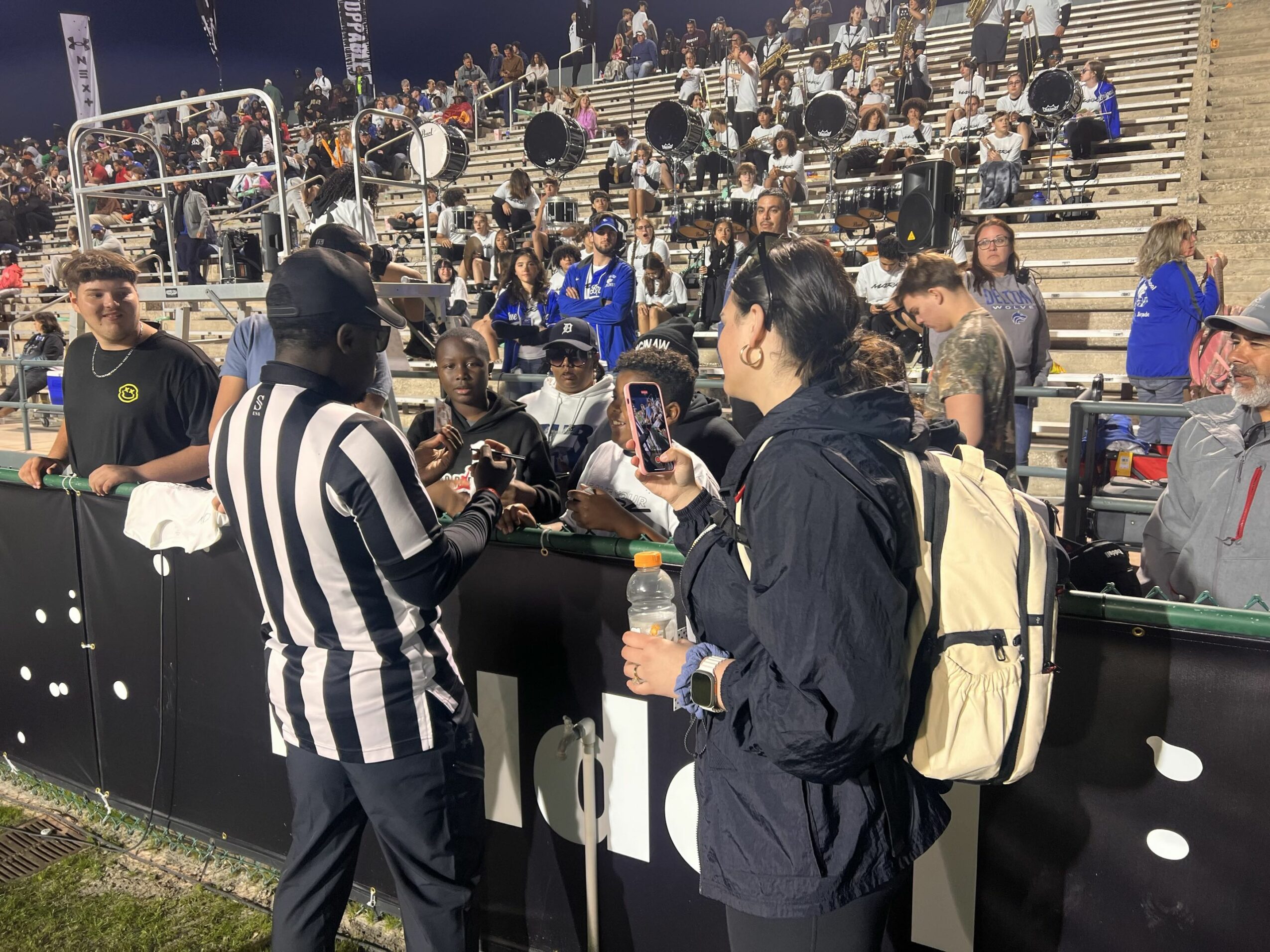 People gather at a sports event, some talking to officials, with stadium seating in the background.