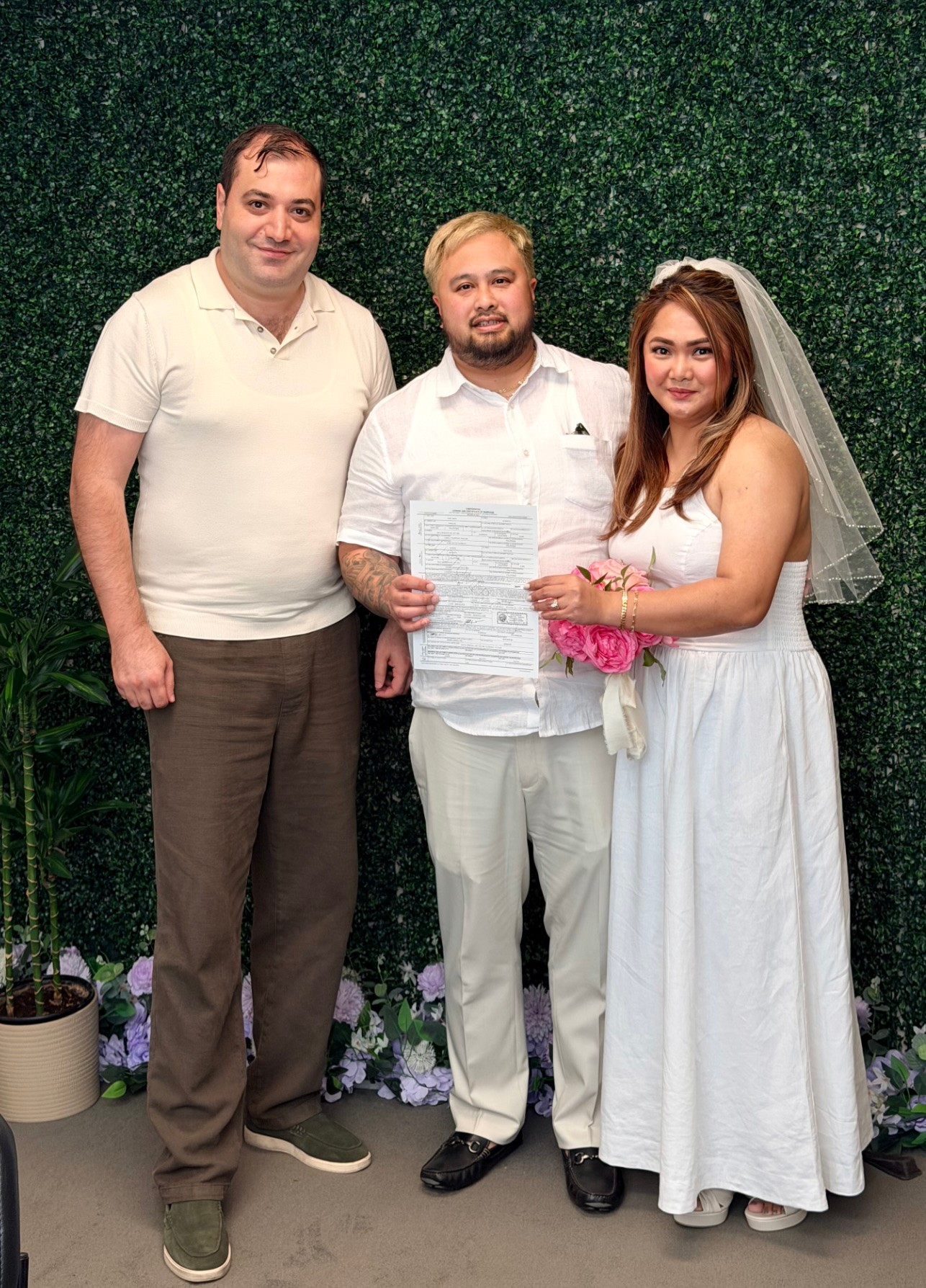 Three people standing in front of a green wall, one woman in a wedding dress and veil, two men in casual and formal attire.