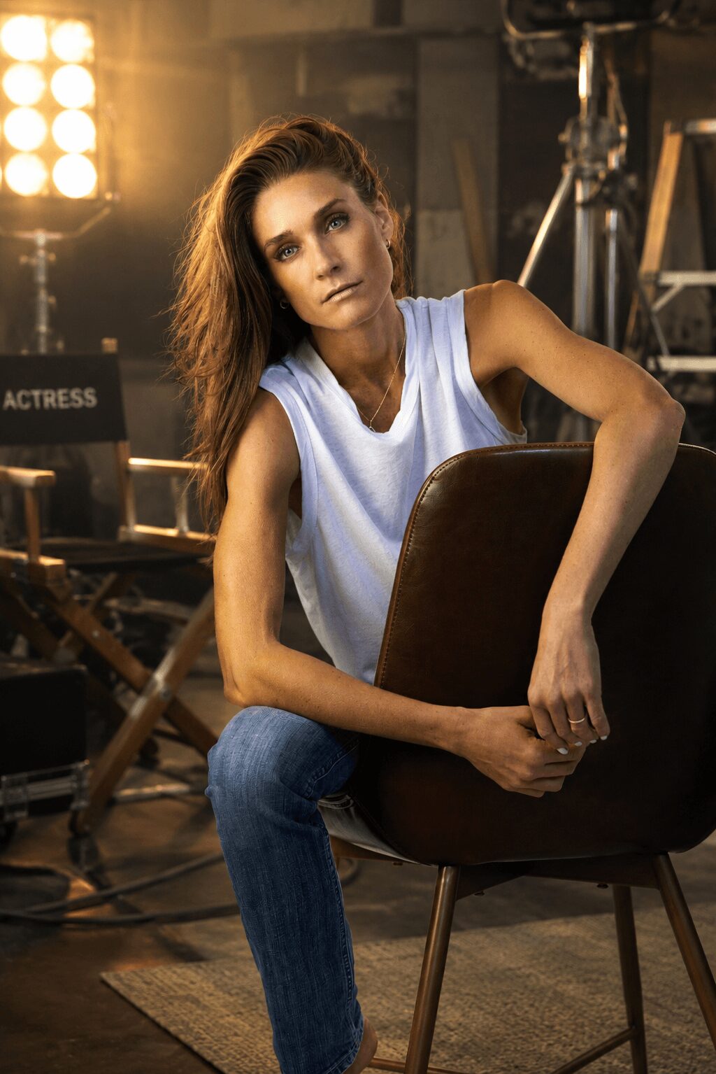 Woman with long hair sitting on a chair in a studio setting, holding the backrest, with professional lighting and equipment around.