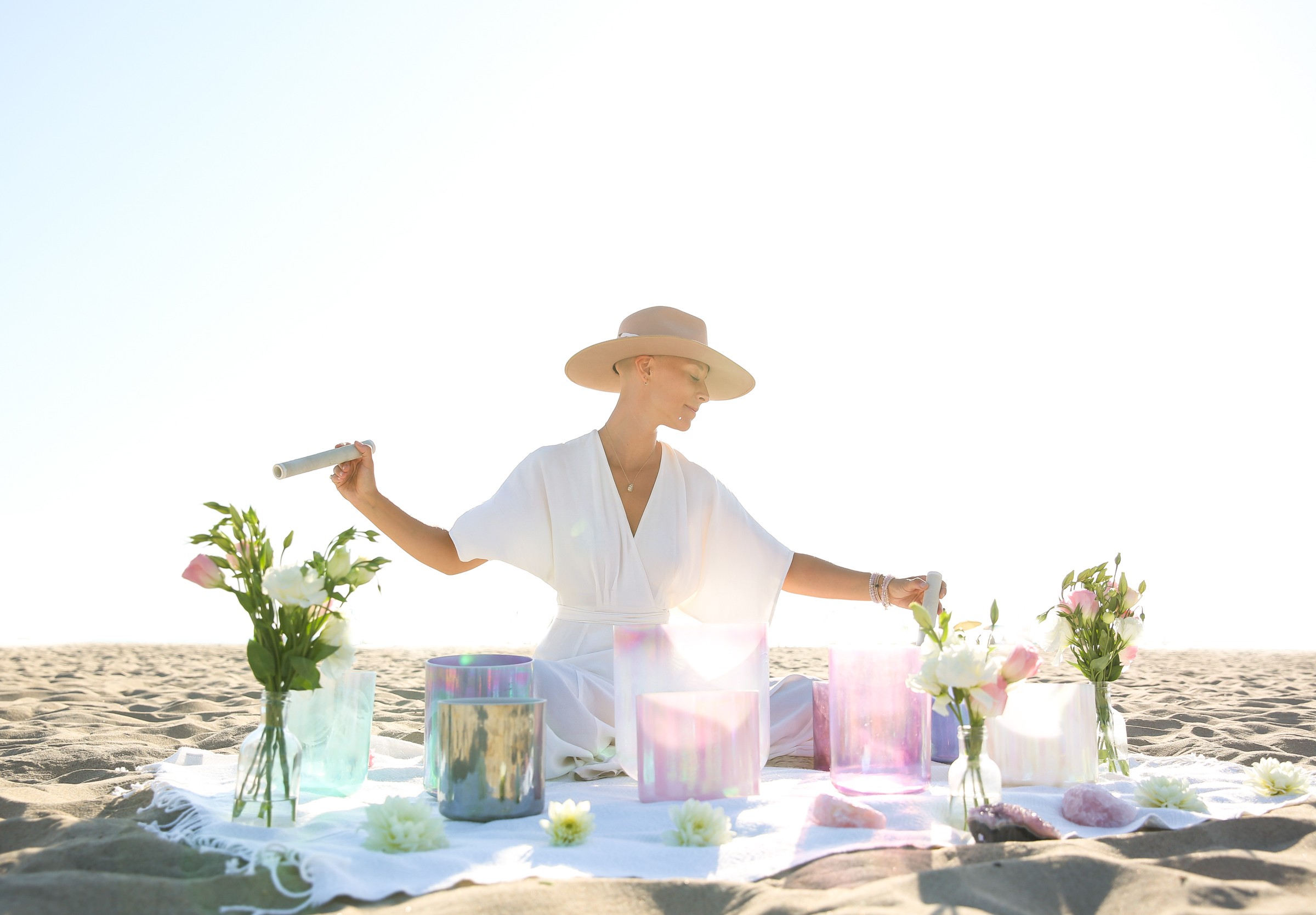 Person wearing a wide-brimmed hat and white dress arranging flowers on a beach, with a table and gift boxes, under bright sky.