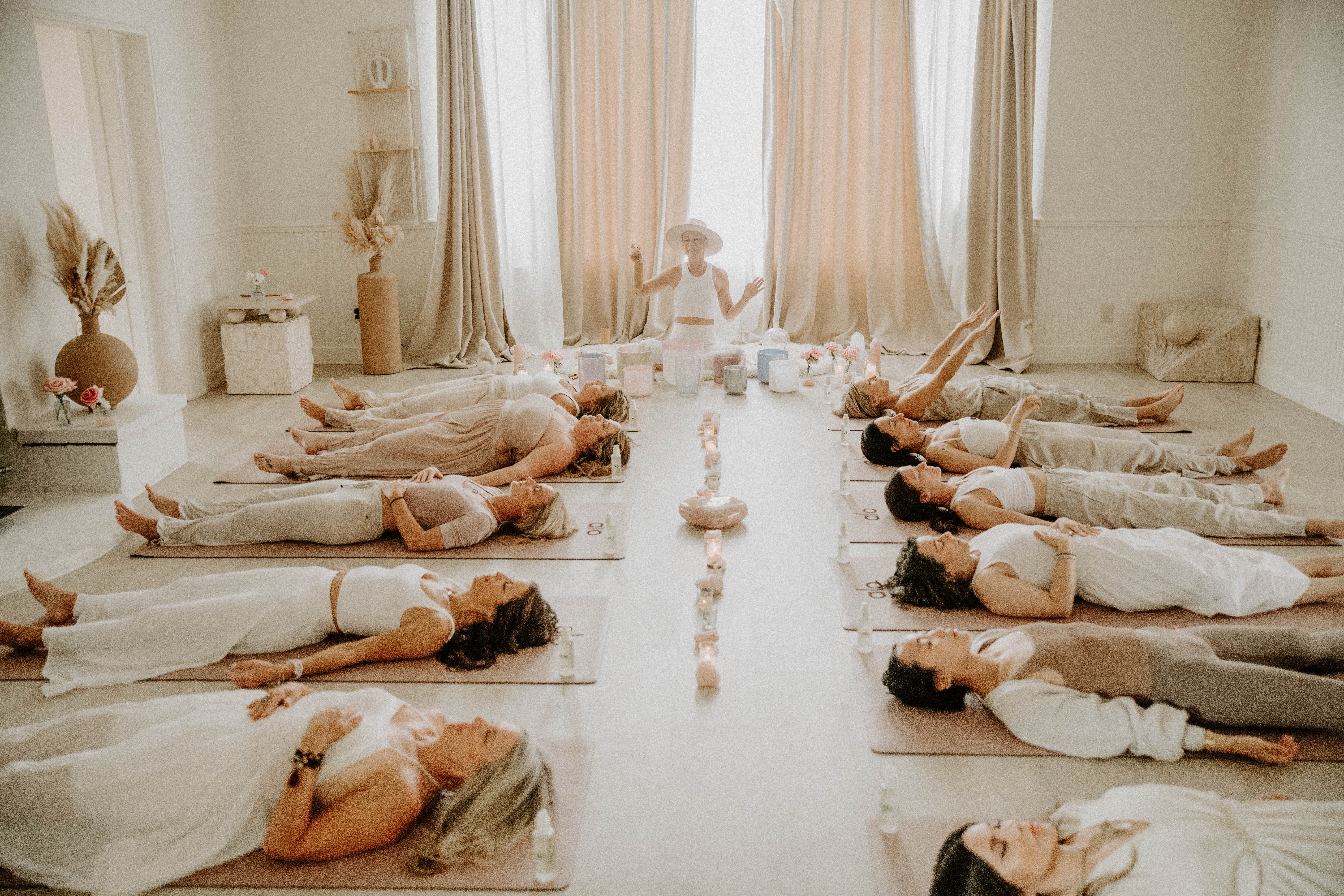 People lying on yoga mats in a bright, airy room with large windows and natural light, practicing meditation or relaxation.