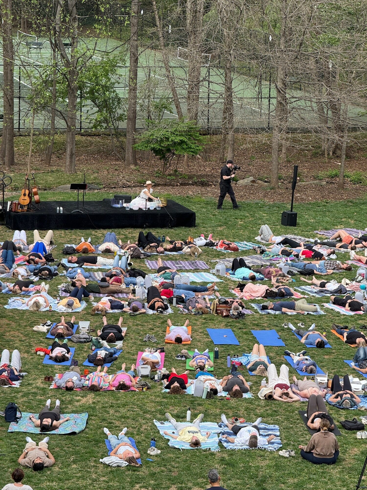 Large outdoor gathering with many people lying on mats and blankets on grass, a stage with performers, and trees in background.