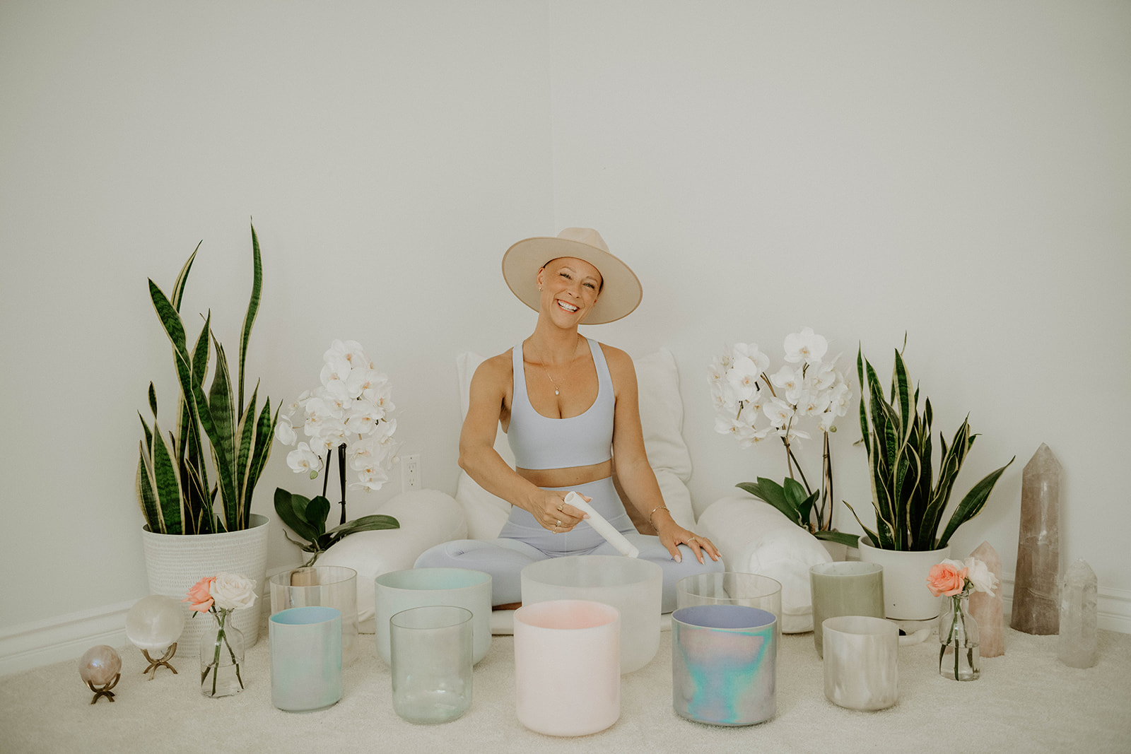 Woman sitting cross-legged on the floor, smiling, wearing a hat and white outfit, surrounded by potted plants and candles.