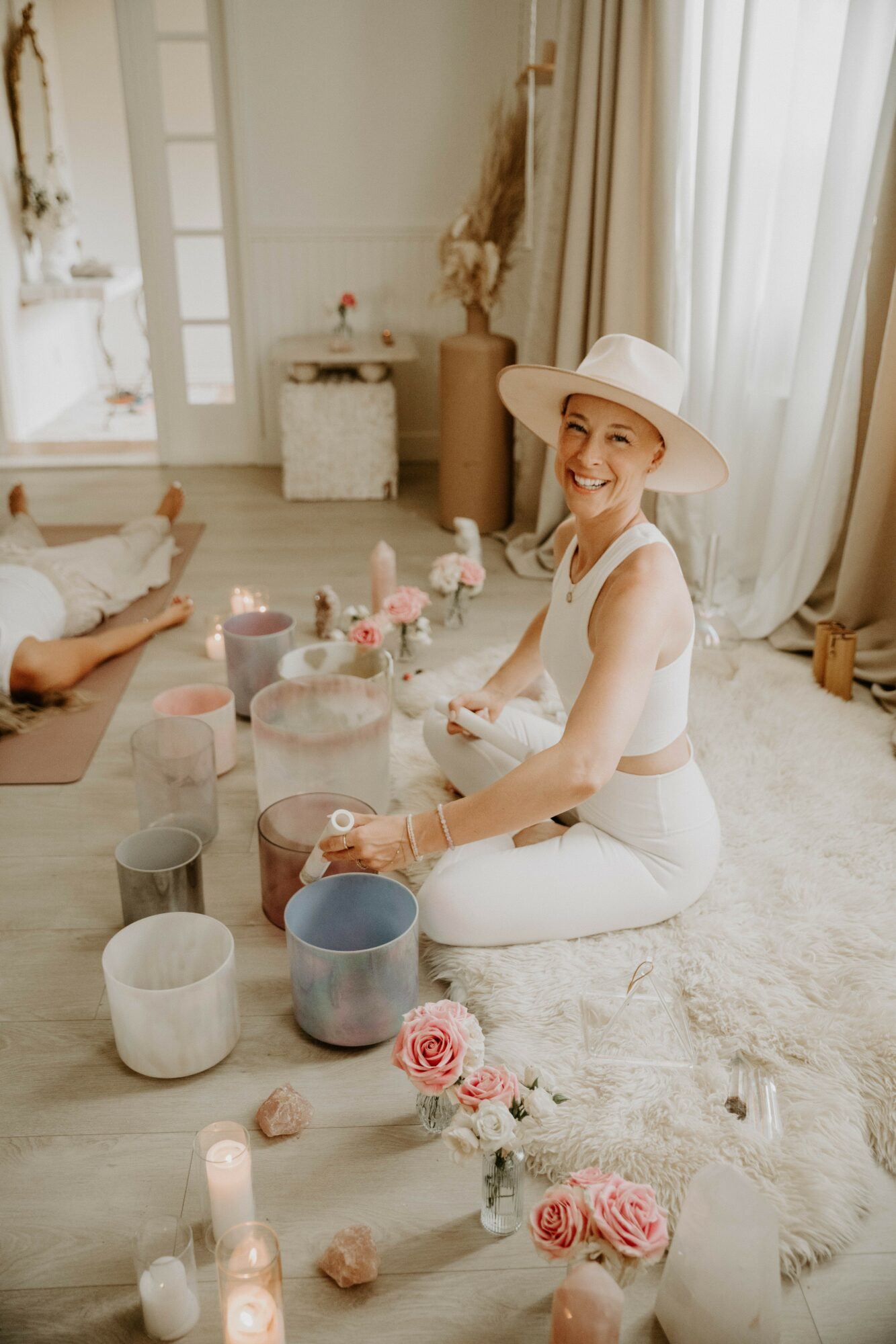 Woman in white hat and outfit sitting on a rug with candles, flowers, and people in a bright room.