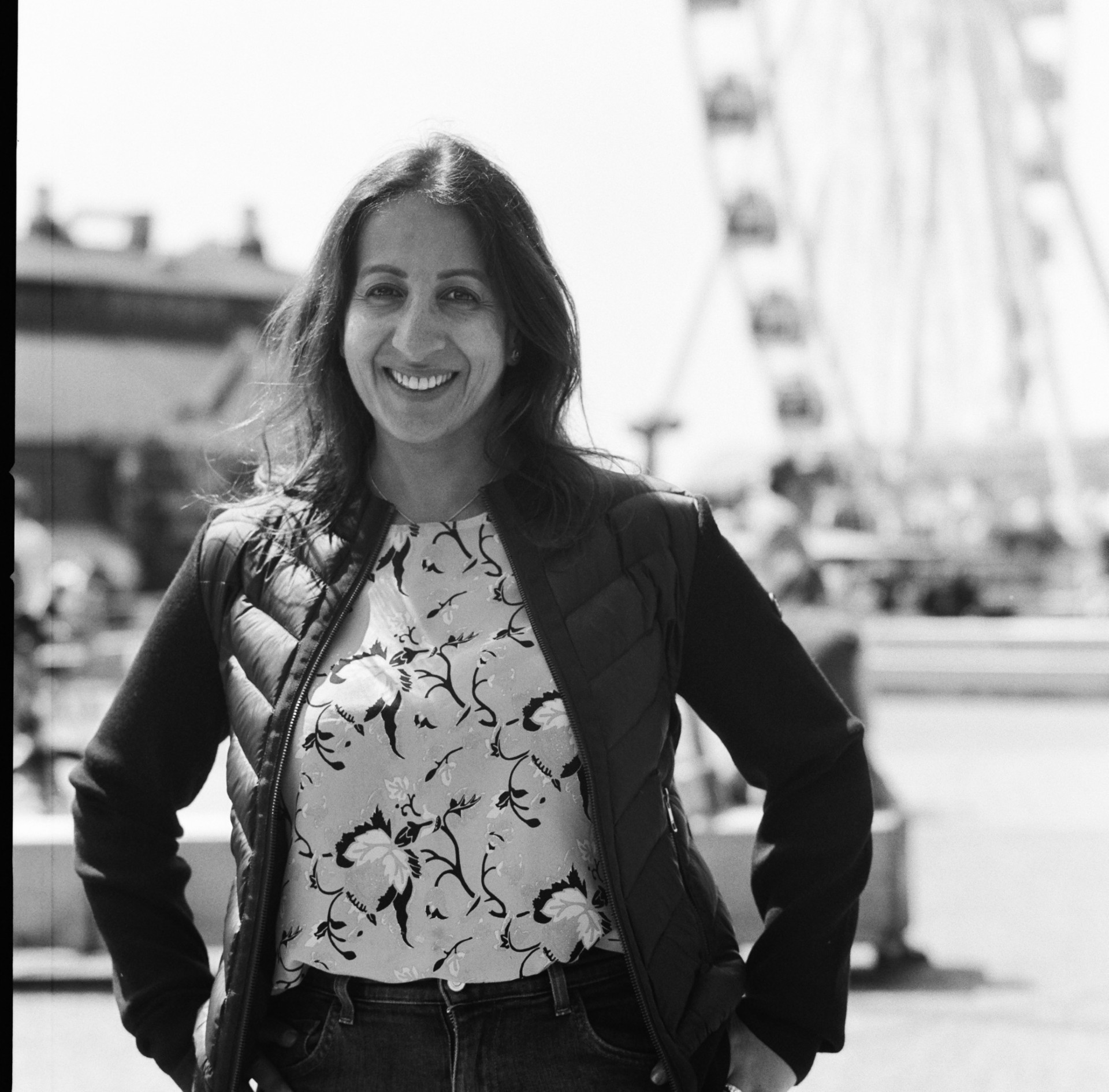 Woman smiling outdoors with a ferris wheel in the background, wearing a patterned top and jacket.