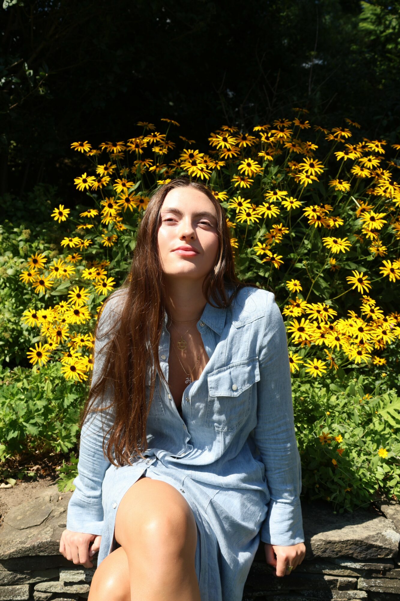 Young woman with long hair sitting outdoors in front of yellow flowers, wearing a light blue dress, sunlight on her face.