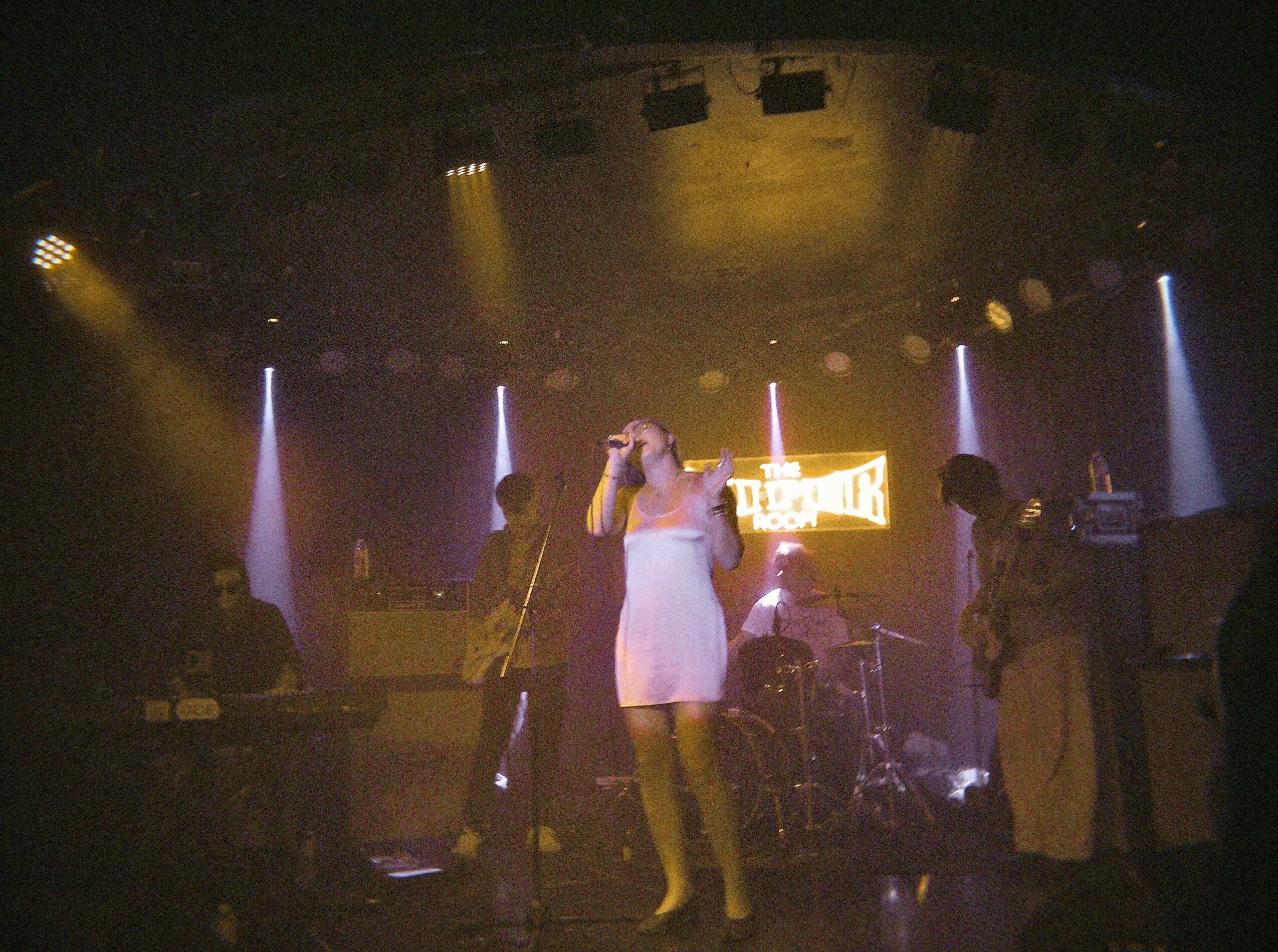 A woman in a white dress sings on stage with a band, colorful lights, and a sign in the background.