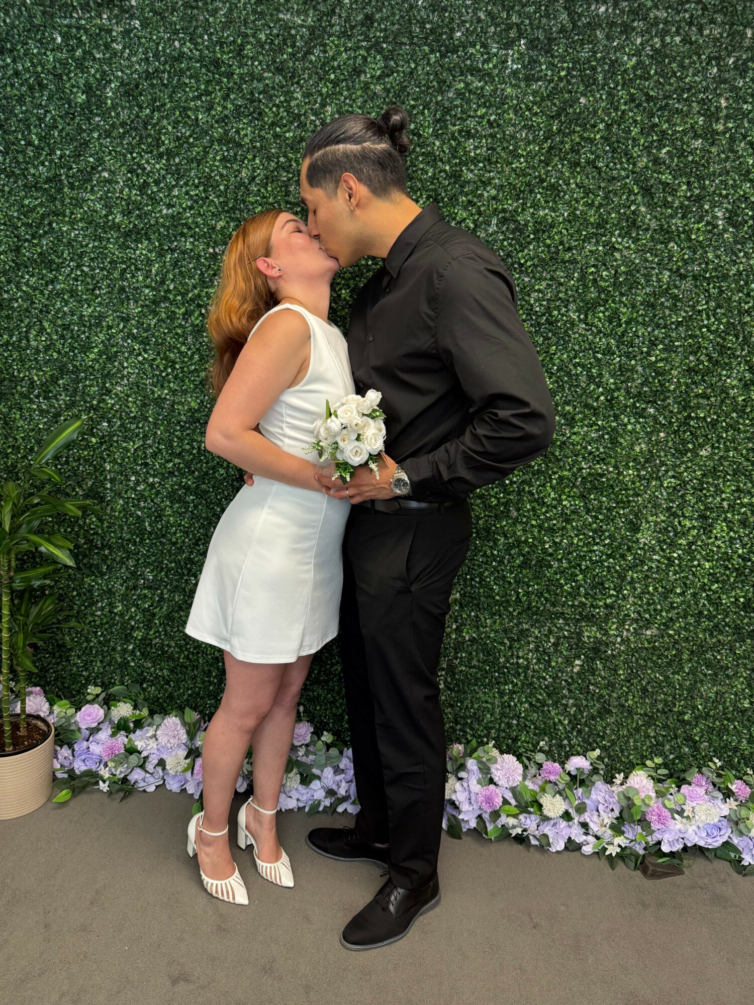 A couple kissing outdoors, woman in white dress holding a bouquet, man in black suit, standing on a flower-lined path.