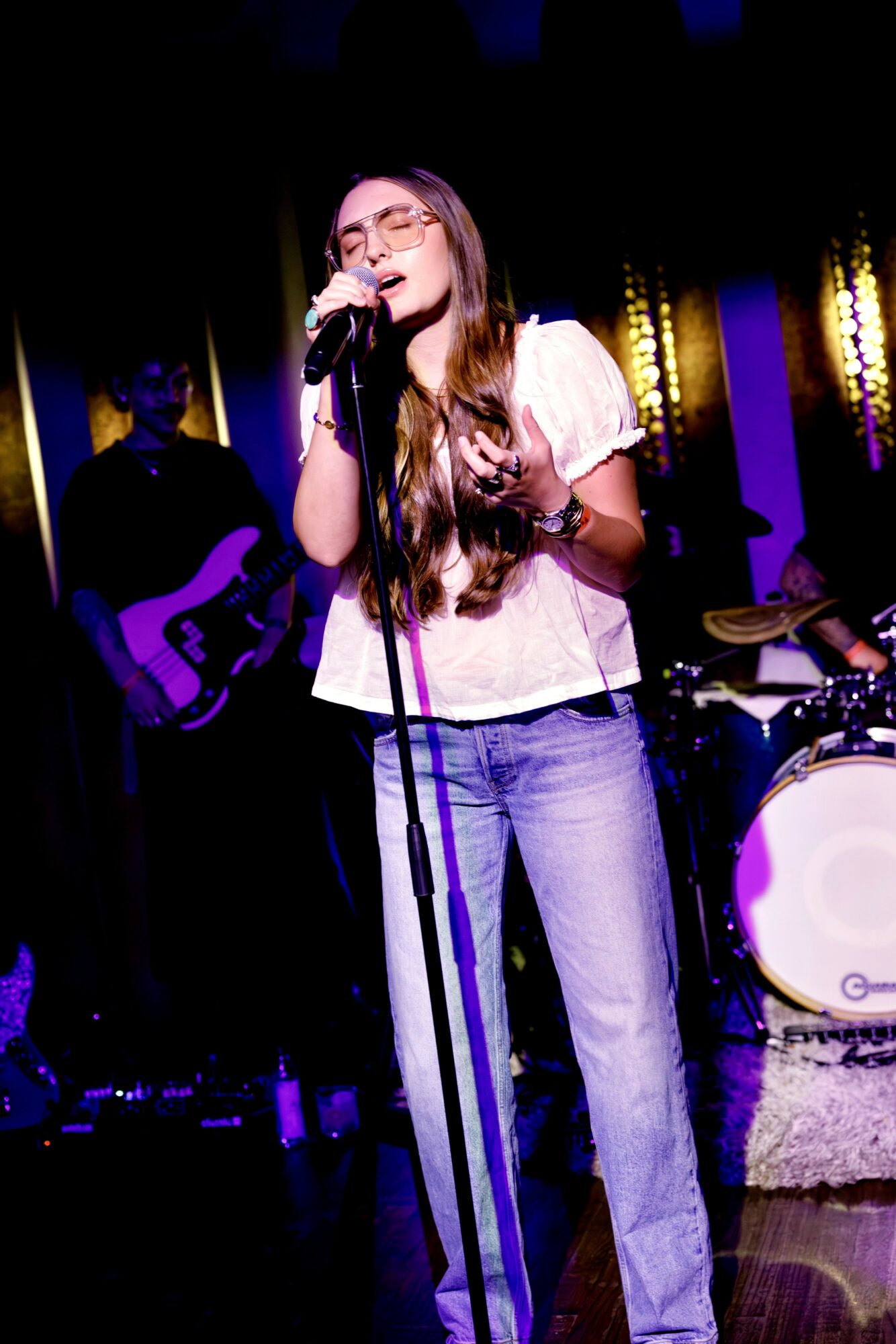 Young woman singing into a microphone on stage with band members playing instruments behind her.