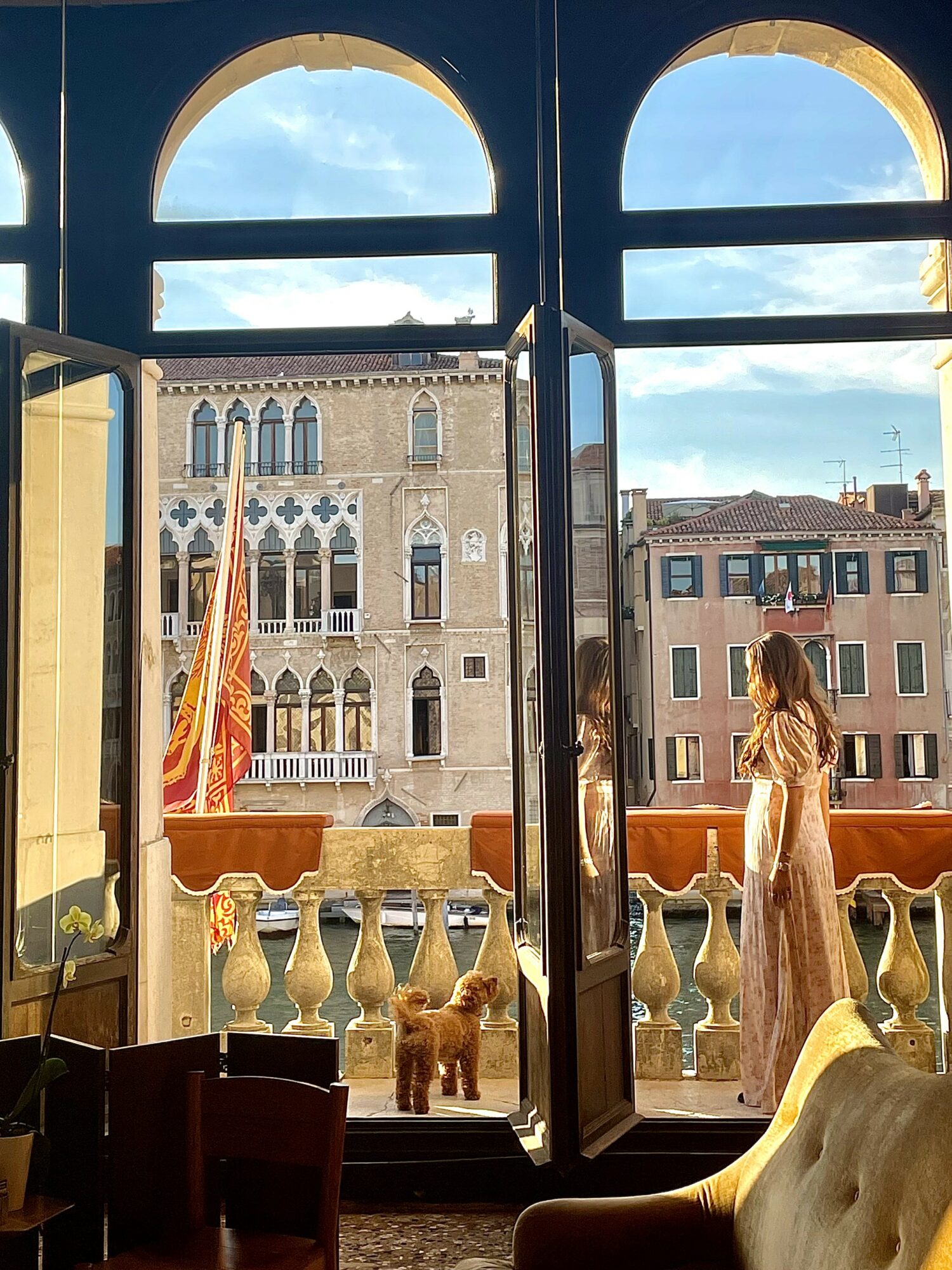 View through open balcony doors showing historic building, woman in dress, and dog outside on balcony, with blue sky and clouds.