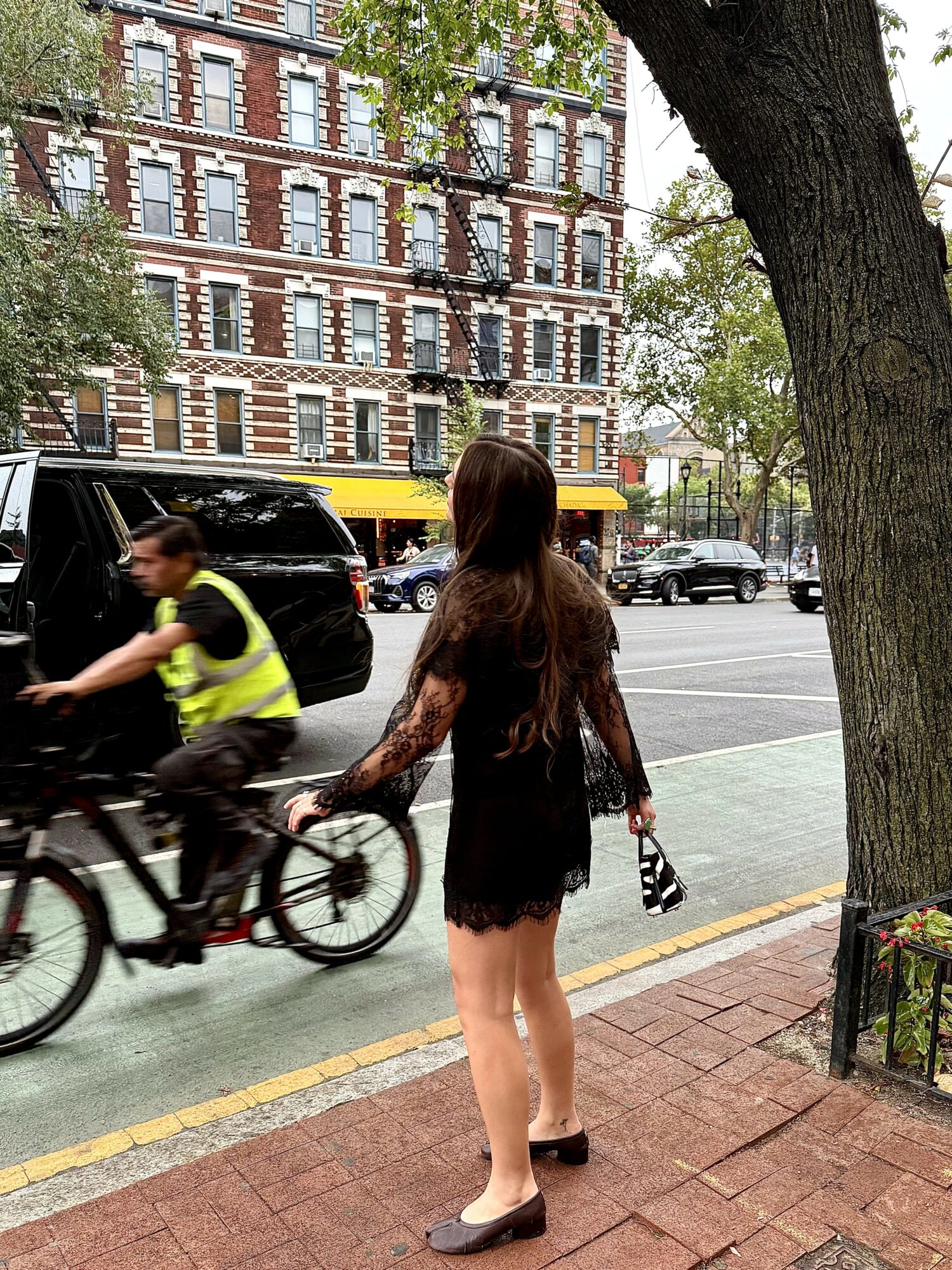Young woman with long hair in a black dress and heels stands on sidewalk, holding a small bag, with city street and trees behind.