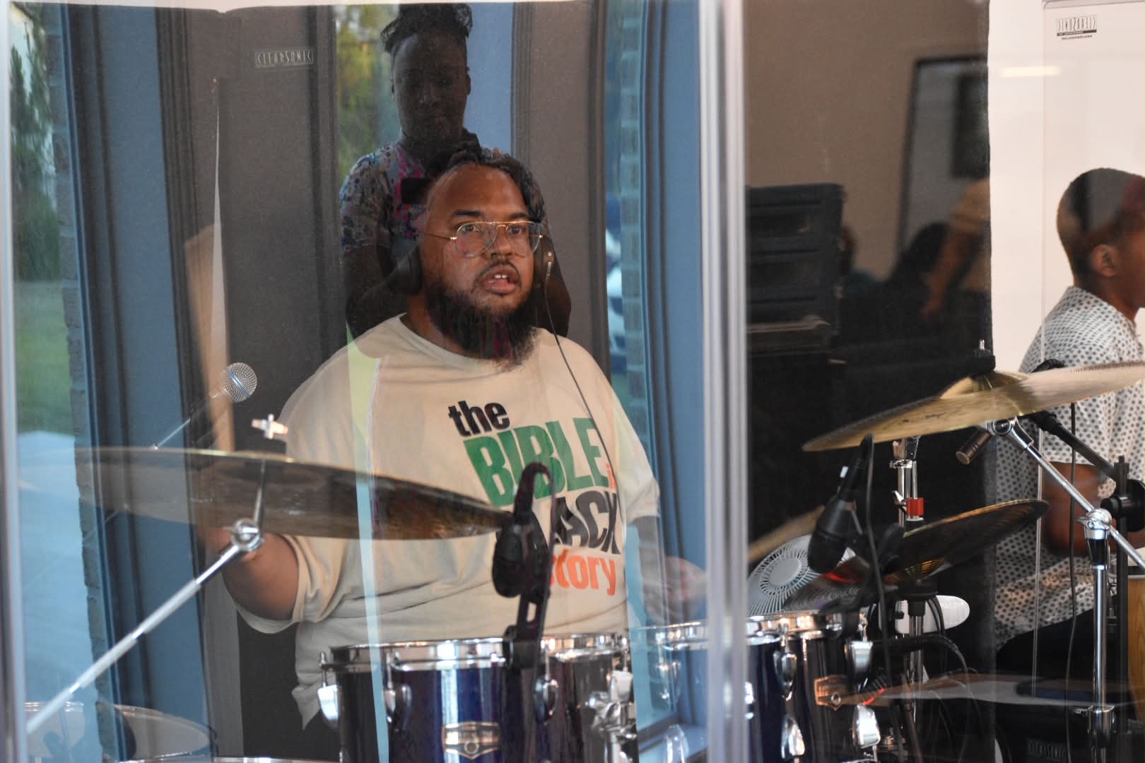 Person with glasses and beard playing drums in a room with glass windows and other people present.