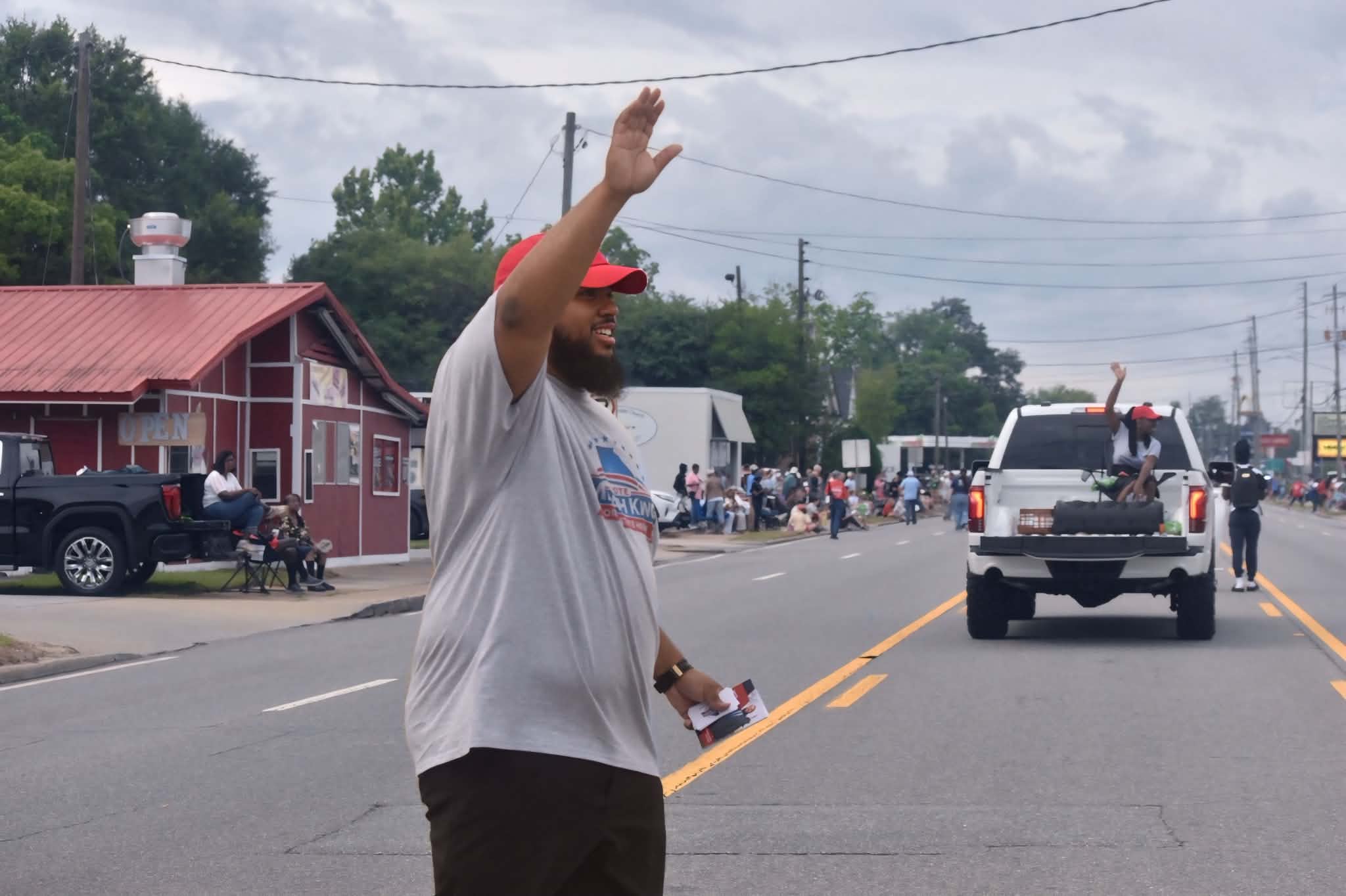 Person with beard and red cap waving on a street with vehicles and buildings in background.