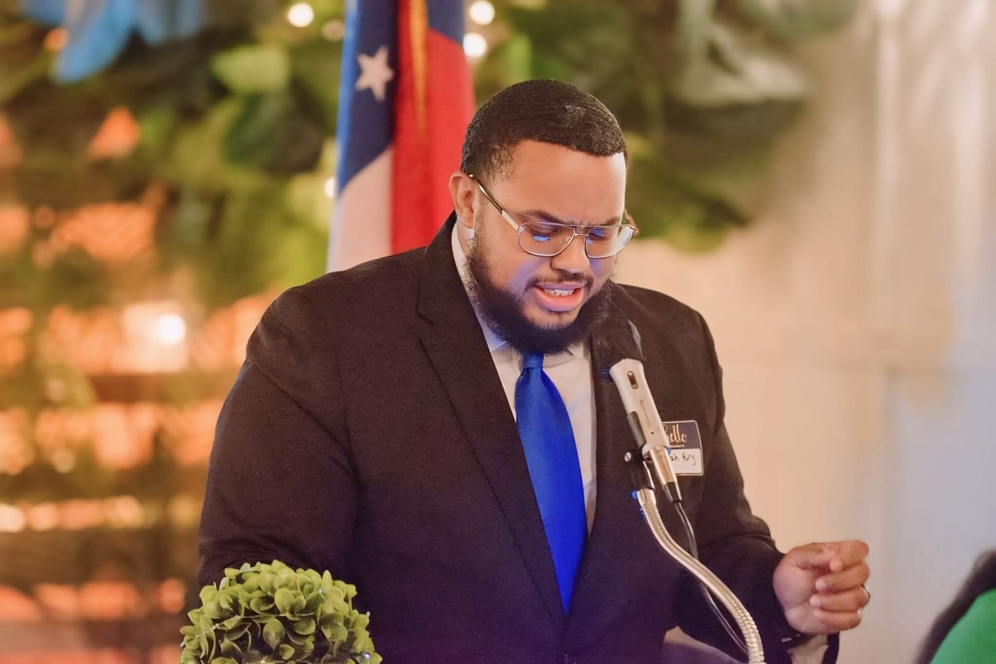 Man in a suit with a blue tie speaking at a microphone, with a flag and greenery in the background.