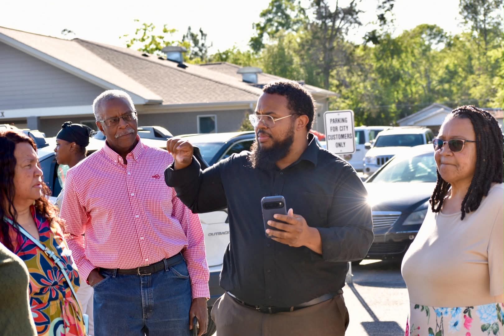 Group of four people talking outdoors in a parking lot, with cars and trees in the background.