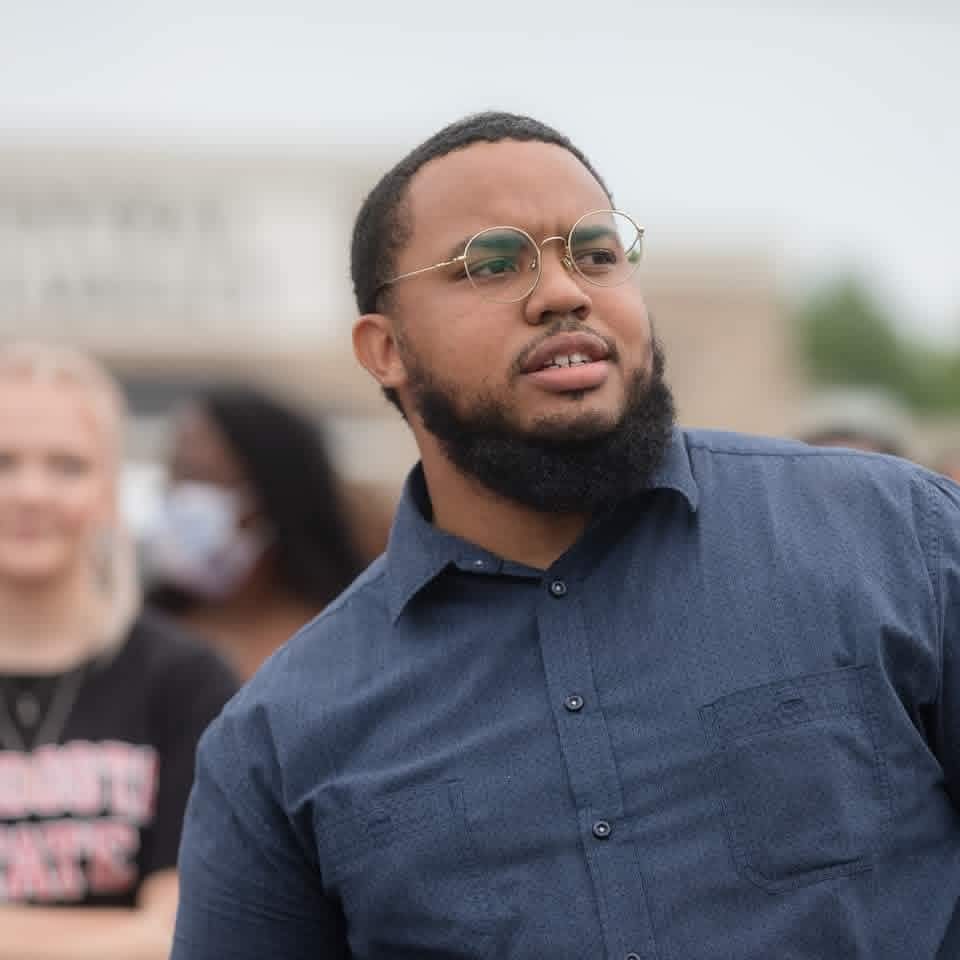 Man with glasses and beard wearing a dark blue shirt outdoors, with blurred people in background.
