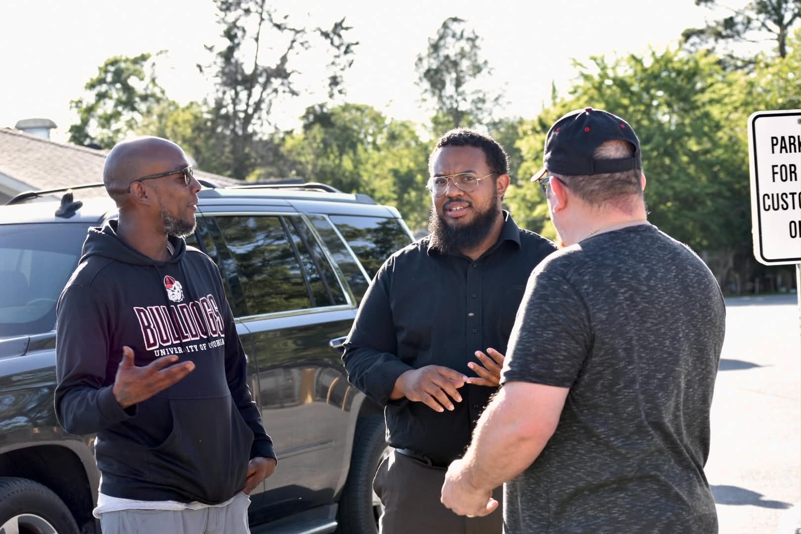 Three men are talking outdoors near a parked car, with trees and a parking sign in the background.