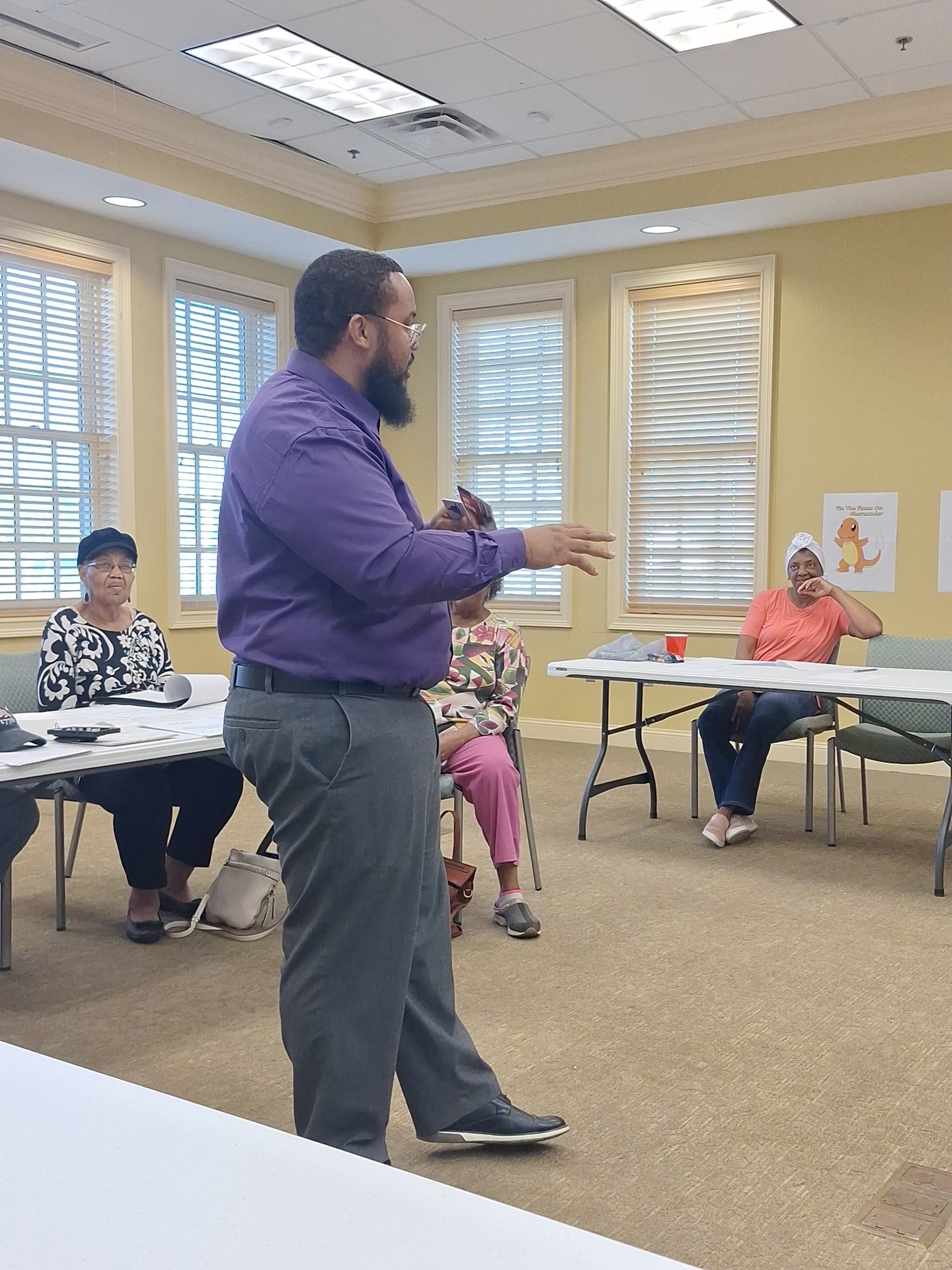Man with beard in purple shirt speaking to group in a room with windows and tables.