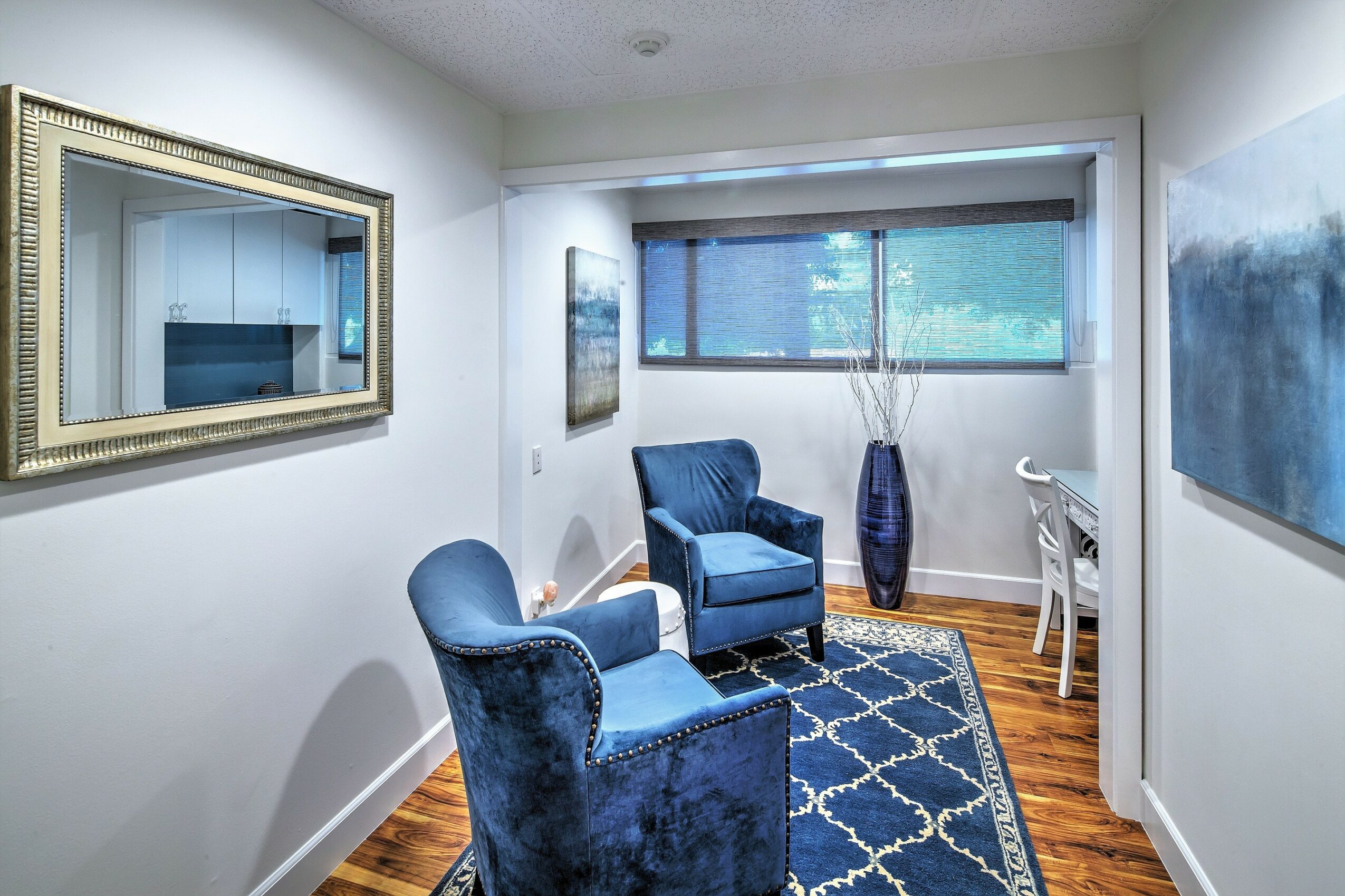 Living room with two blue armchairs, a patterned rug, a large window, and wall art, with a wooden floor.