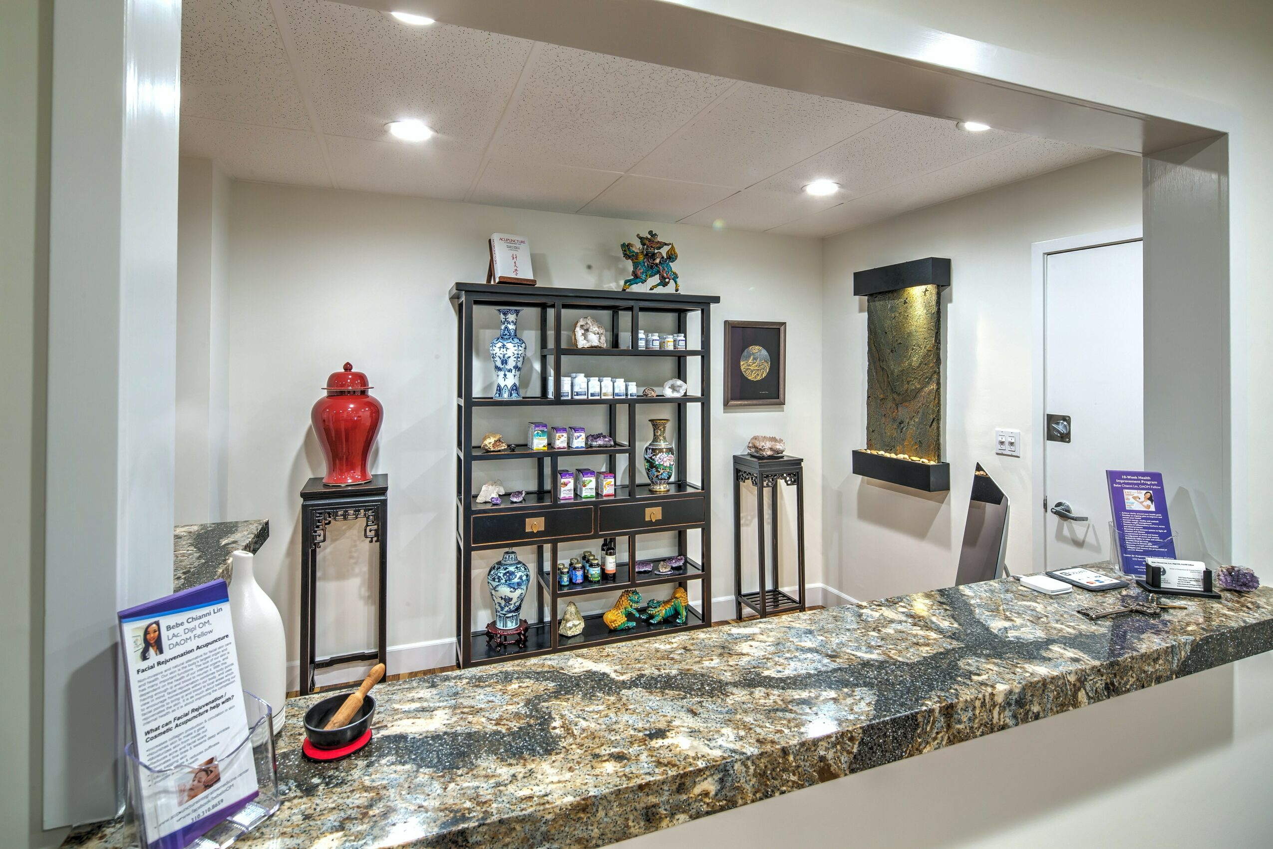 Reception area with a granite counter, black shelving unit, decorative items, and a small table, with a door on the right.