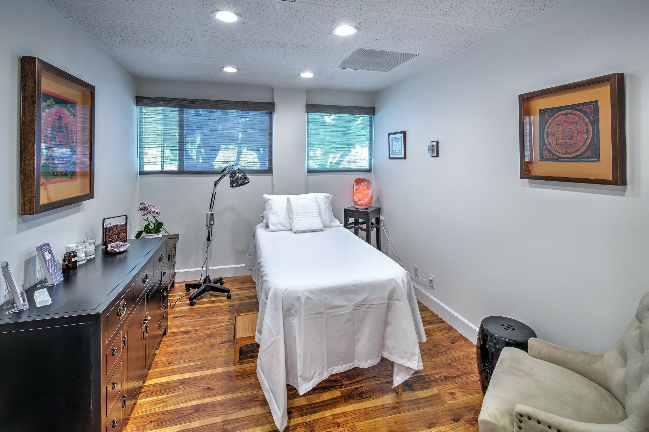 Hospital room with bed, artwork, dresser, and chair, illuminated by natural light from windows, with hardwood floor.