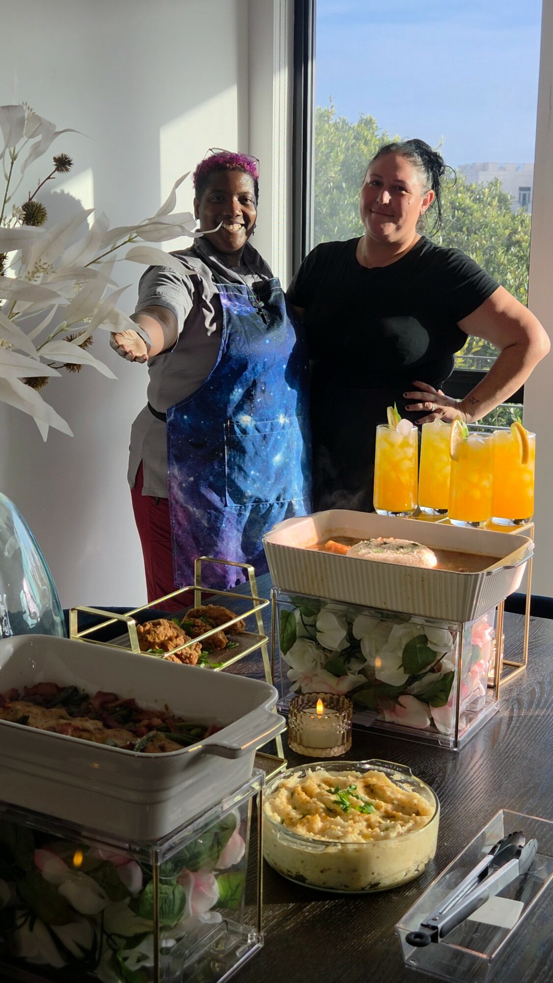 Two women standing behind a table with food and drinks, near a window with a view outside, smiling at the camera.