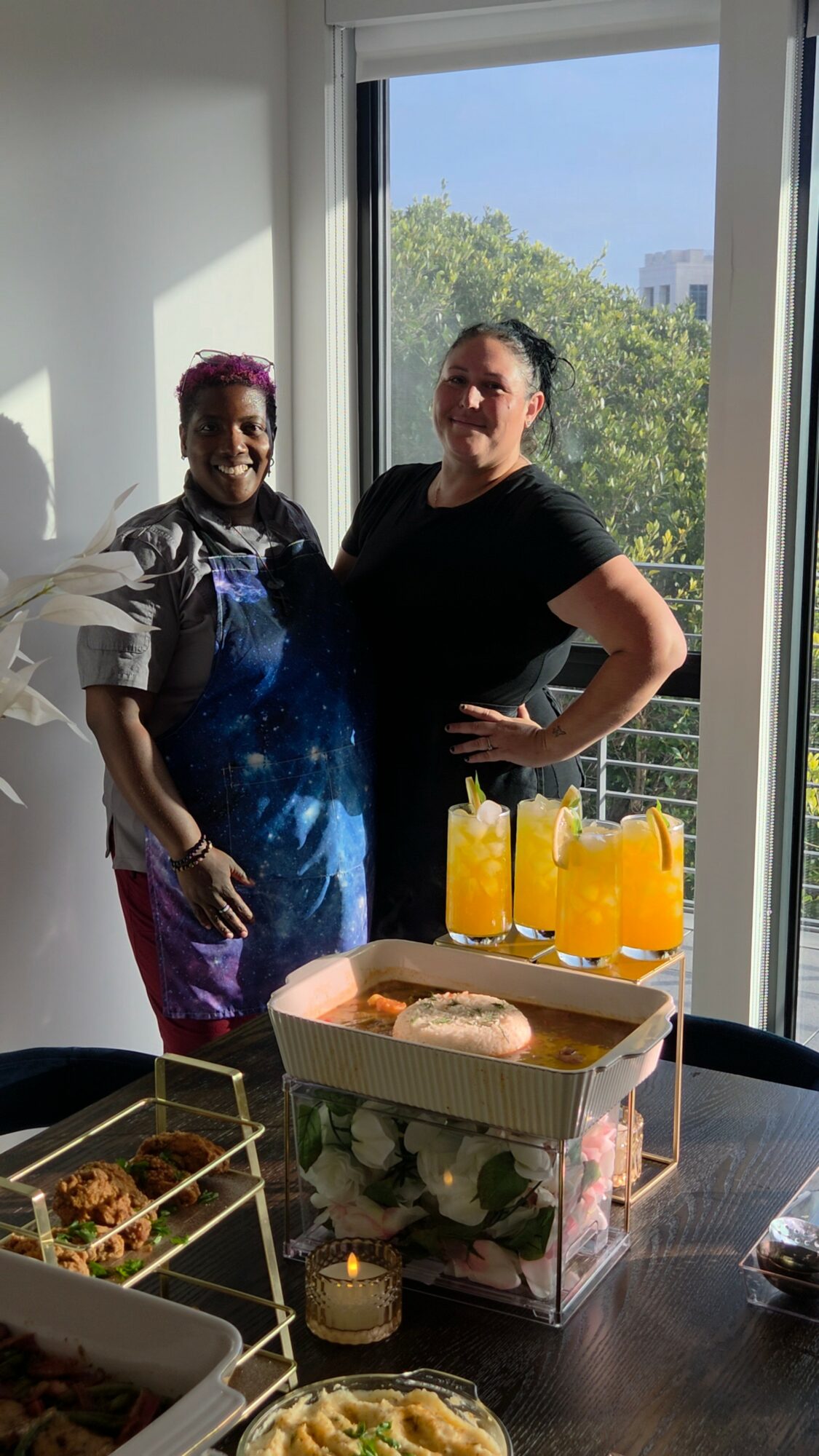 Two women standing indoors near a window with trees and buildings outside, a table with drinks and food in front of them.