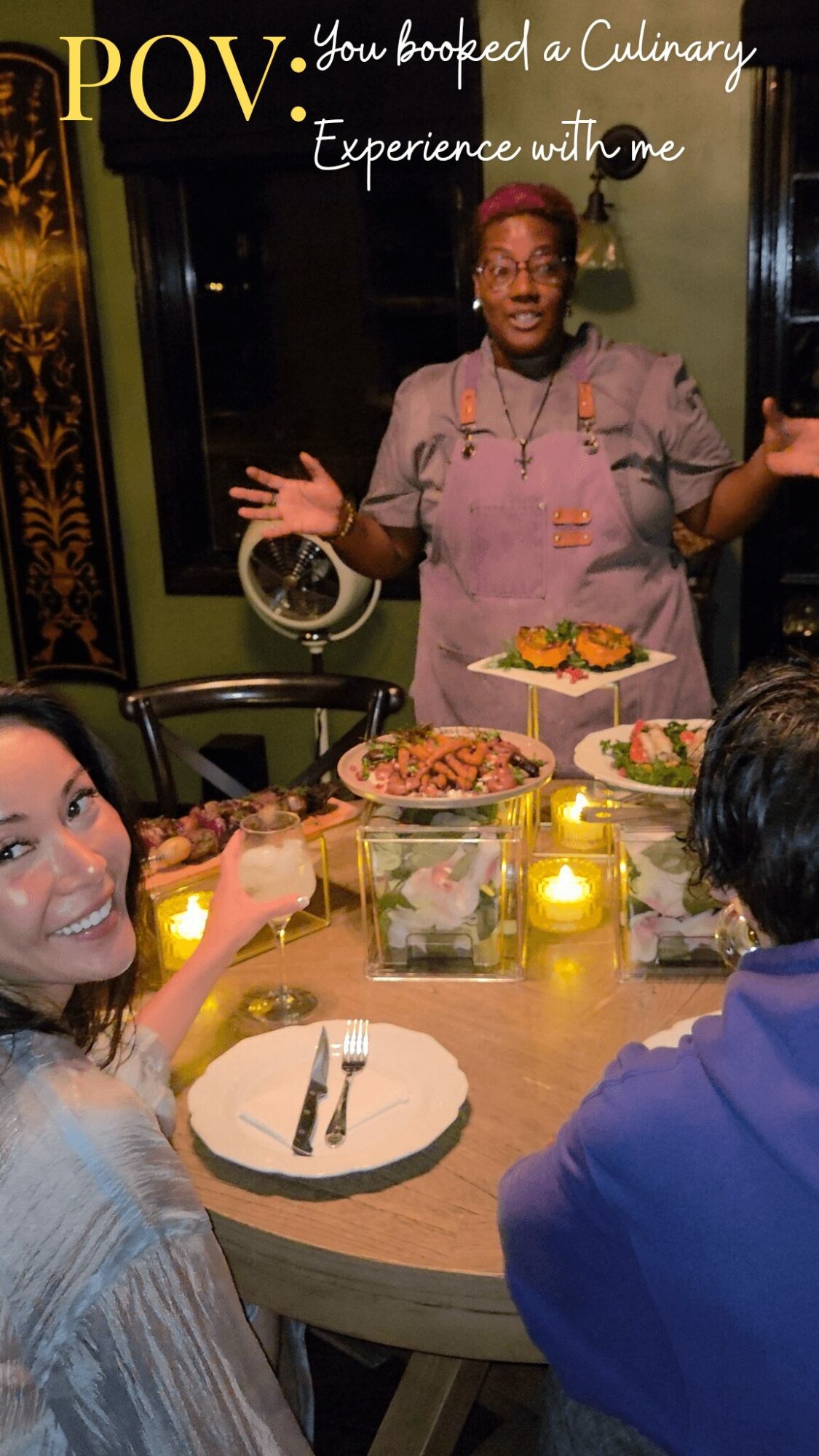 Person in chef's uniform standing behind a dining table with food, two people seated at the table, candles, and a mirror in the background.